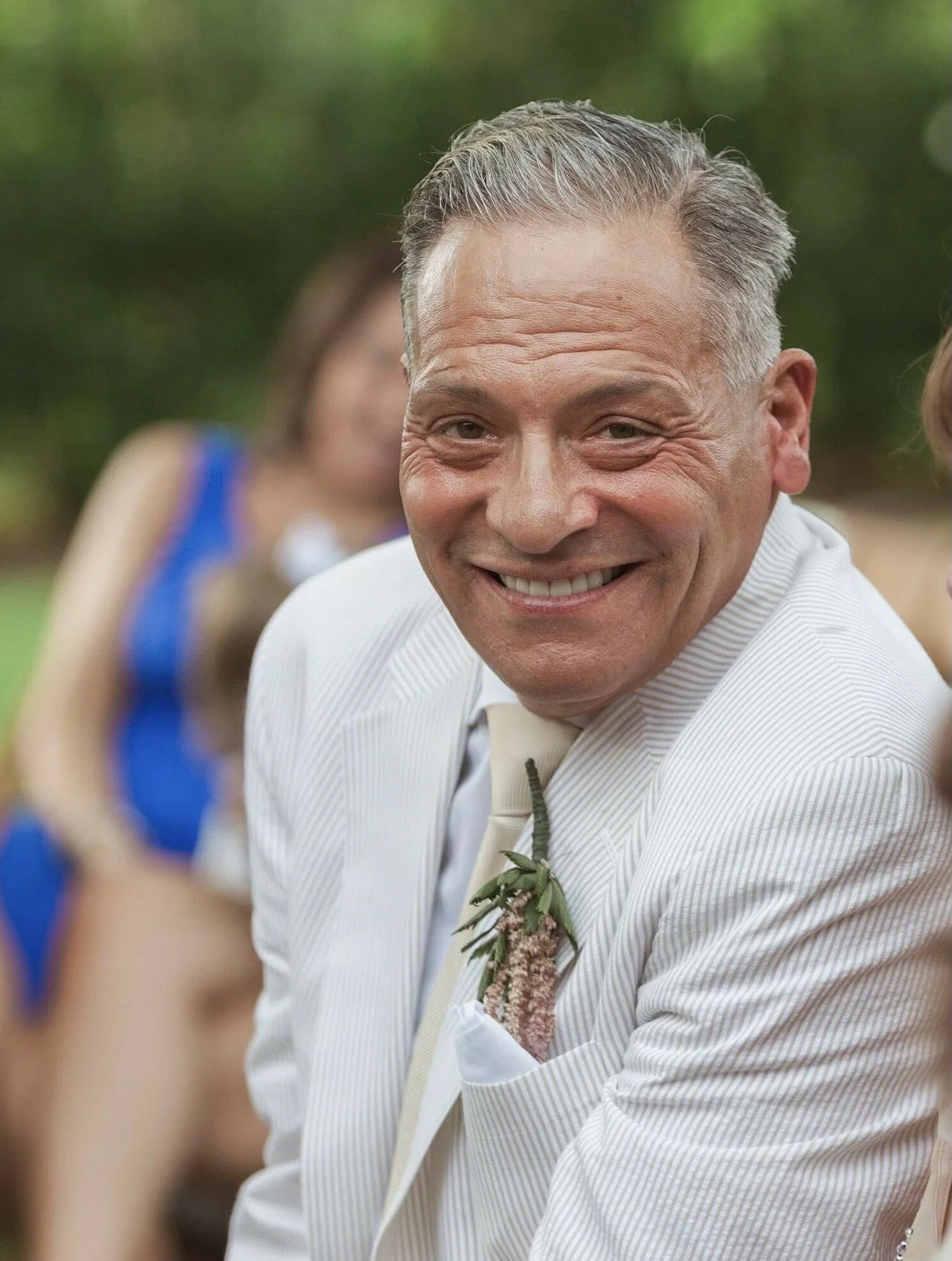 A smiling Perry Stio in a white suit at an outdoor event, with blurred people in colorful outfits in the background.