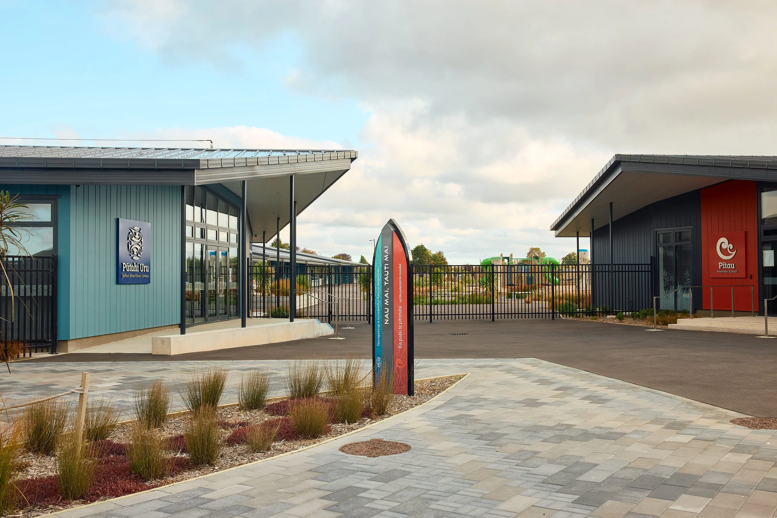 A modern school entrance with two buildings, one painted blue and the other painted black with red accents, separated by a black fence. There are signs indicating school names and a landscaped area with plants and paved walkways.