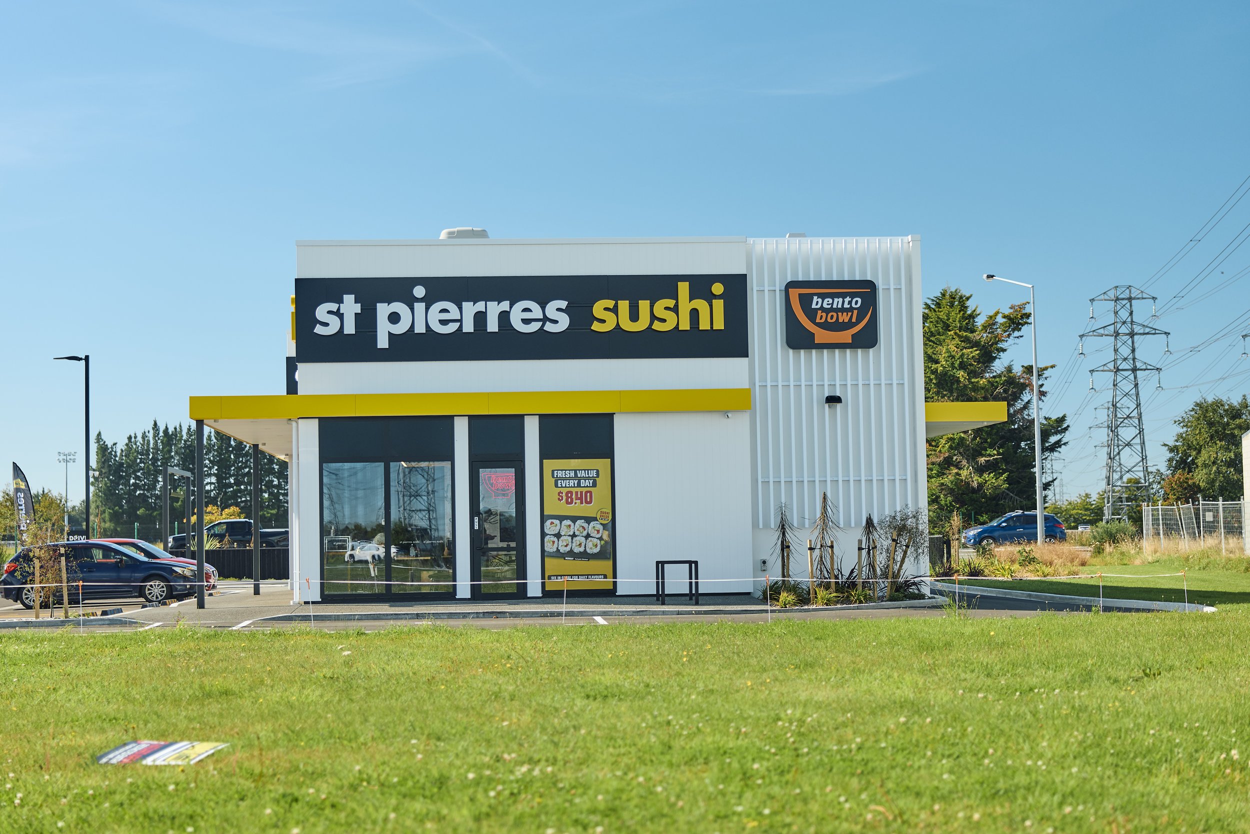 Storefront with a sign reading 'st pierrs sushi' and a smaller sign with a bowl icon labeled 'bento bowl', situated under a clear blue sky with cars in the parking lot and some trees and power lines in the background.