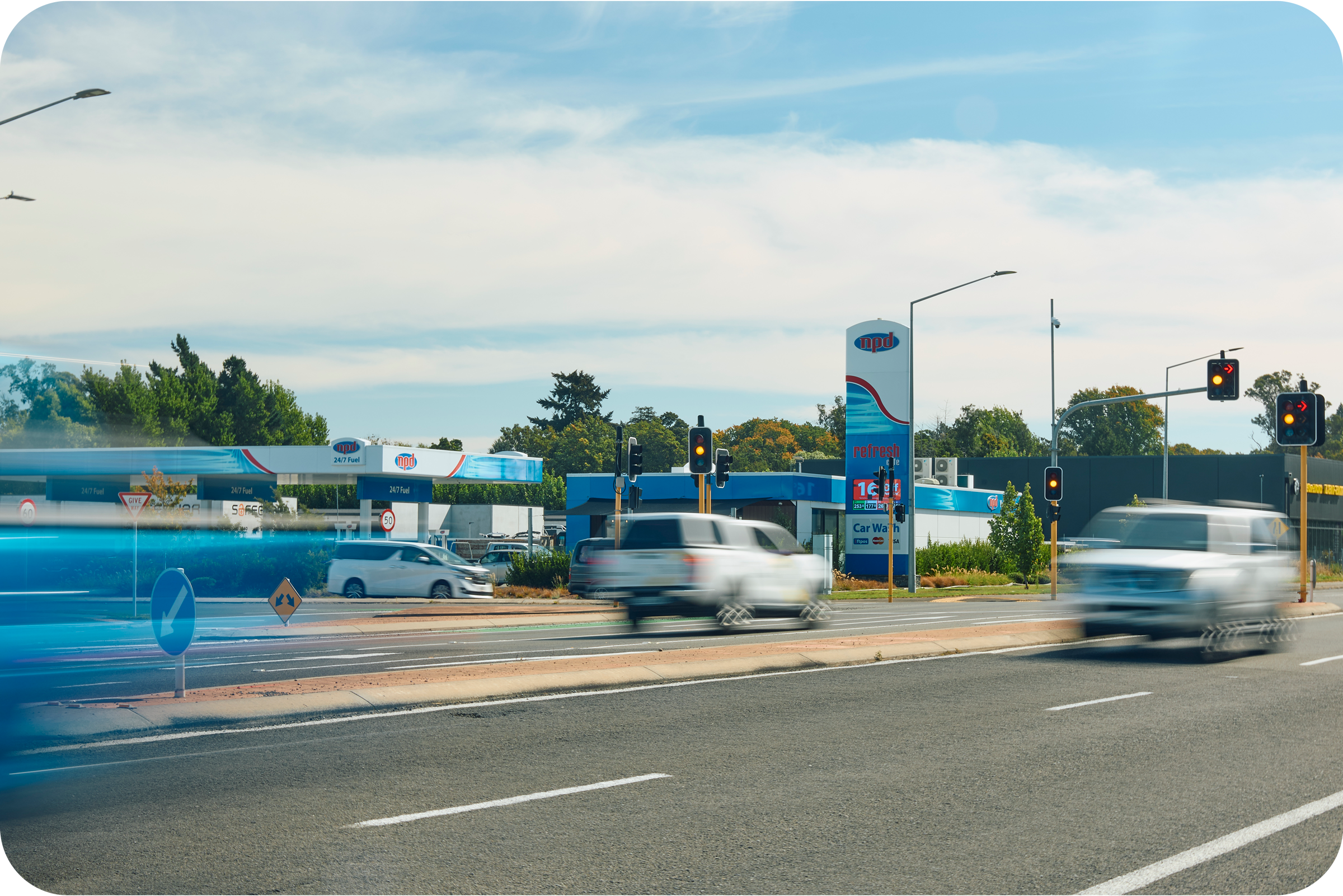 A busy roadside scene with cars driving past a gas station, which has a sign indicating 24/7 fuel, and traffic lights showing red and amber signals. There are trees in the background and a partly cloudy sky overhead.
