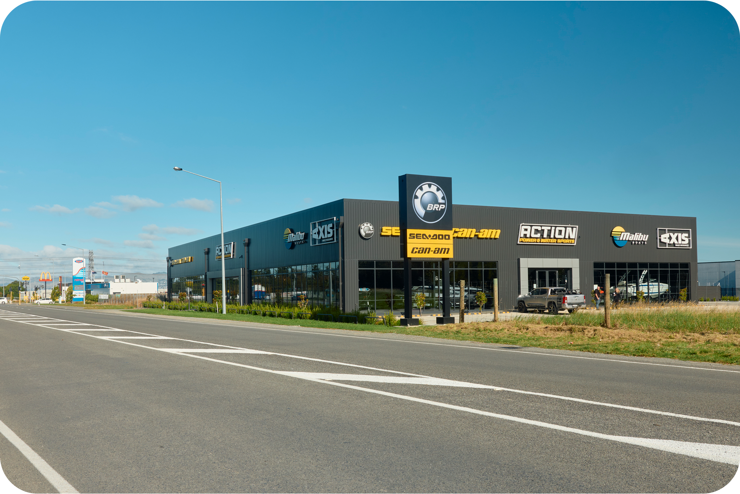 Exterior of a business store with various brand logos, including BRP, Sea-Doo, Can-Am, Malibu Boats, Axis, and Action Power & Water Sports, near a main road with a clear blue sky.