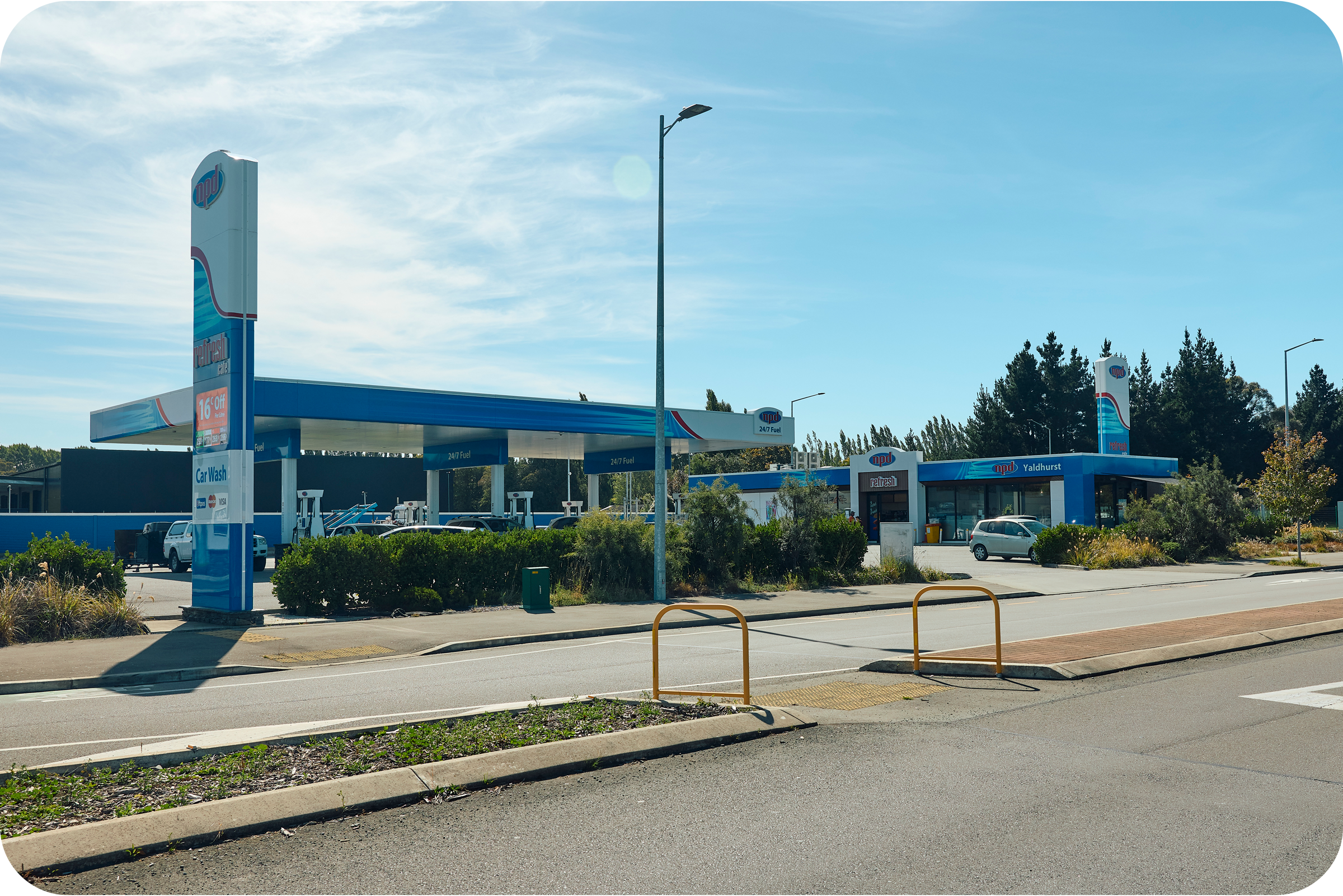 A Shell gas station with multiple fuel pumps, a convenience store, and parking area, located along a street with a strip of bushes and a sidewalk in the foreground. There are trees in the background under a partly cloudy sky.