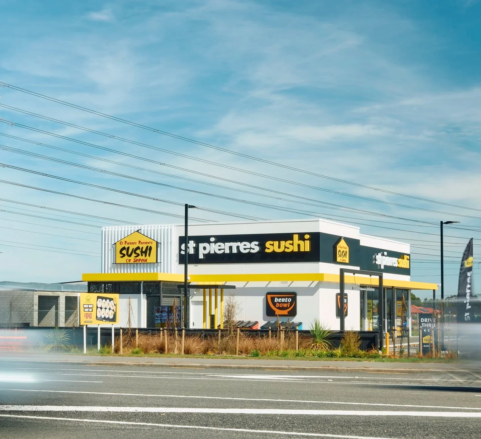 An exterior view of a sushi restaurant called St Pierres, with signage indicating it offers Japanese sushi and bento bowls, under a clear blue sky with power lines overhead.