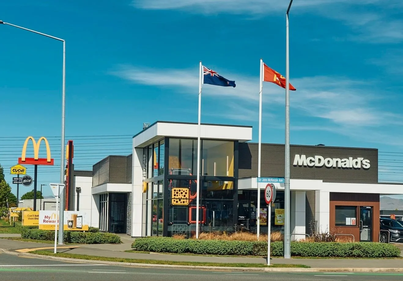 Front view of a McDonald's restaurant with flags of New Zealand and Australia flying above, a glass entrance, and a McDonald's sign on the building. There are signs near the entrance and parked cars nearby.