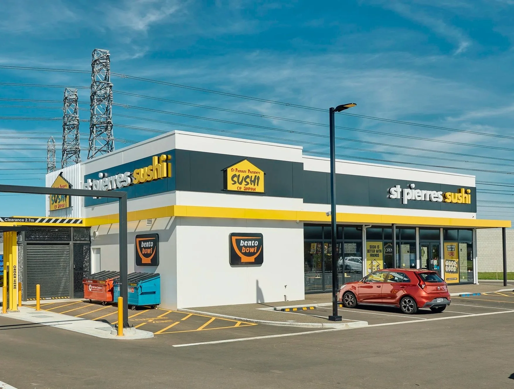 A shopping plaza with a building that has signs for St. Pierres Sushi and Bento Bowl in a large parking lot. There are electric towers in the background and a clear blue sky.