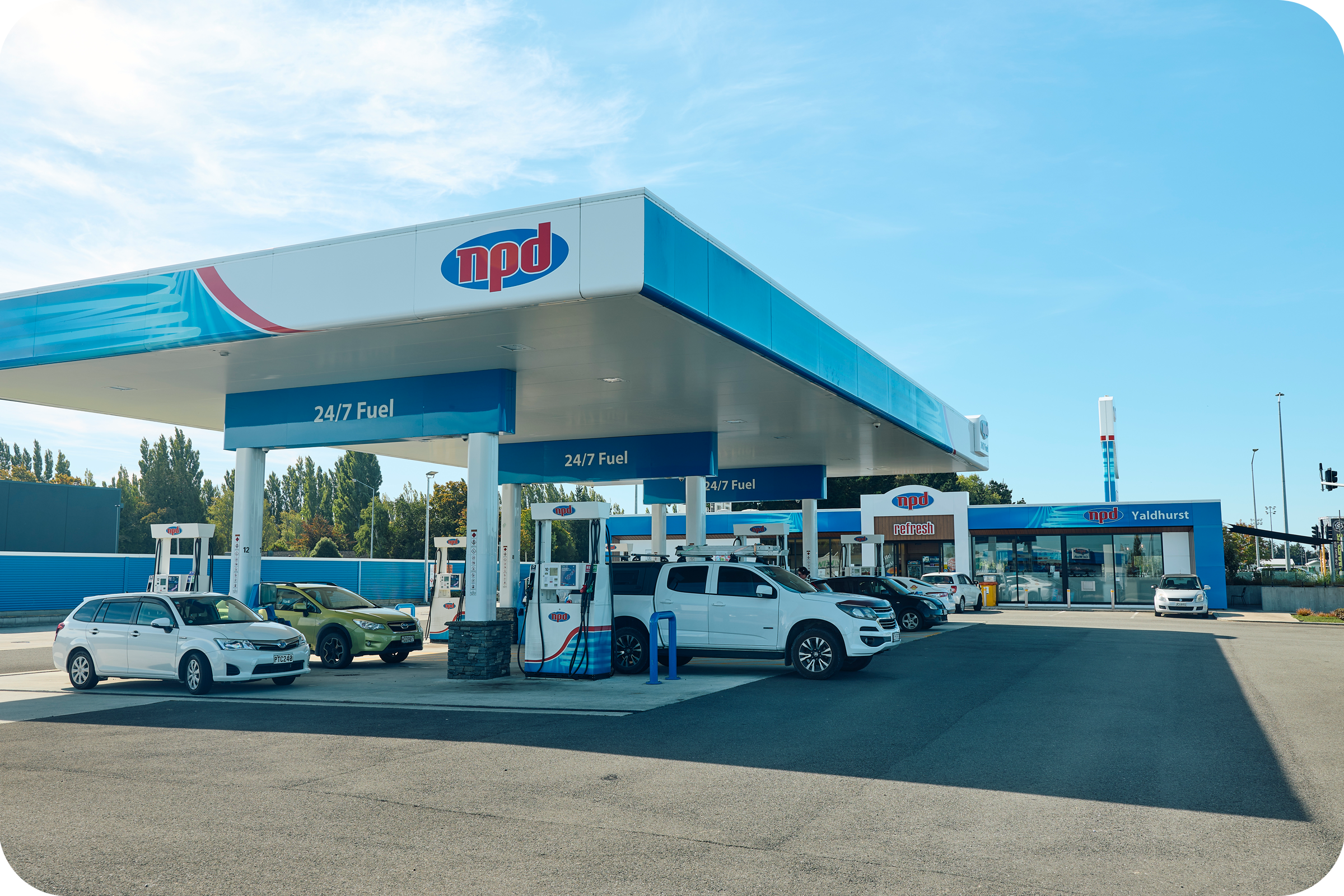 A gas station with the logo 'npd' on the canopy over fueling pumps, several cars parked, and a small convenience store in the background.