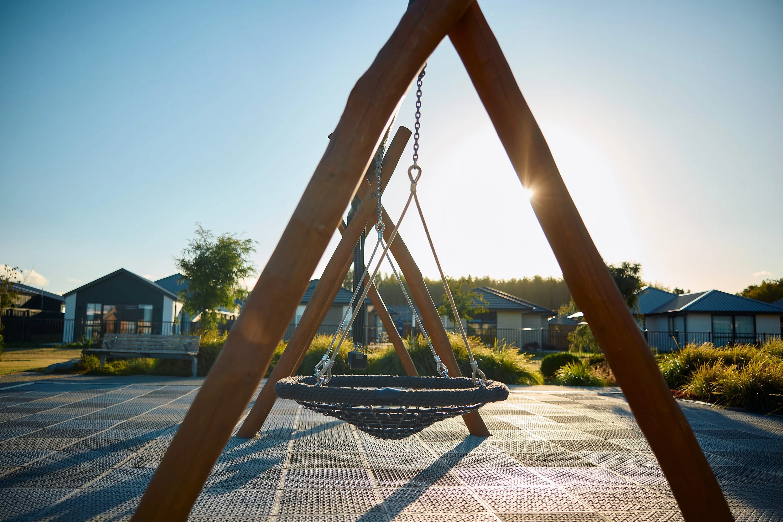Empty swing set in a playground with houses in the background, the sun shining brightly behind it at Belfast subdivision