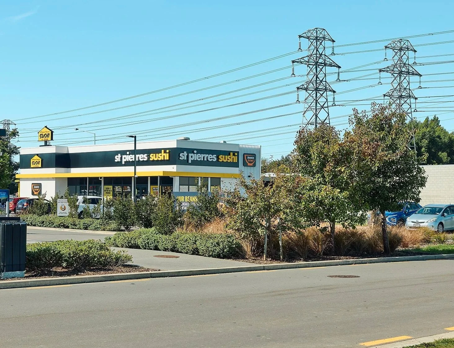 A shopping plaza with a St. Pierre's Sushi restaurant, cars parked and trees in front, power lines above, and clear blue sky.