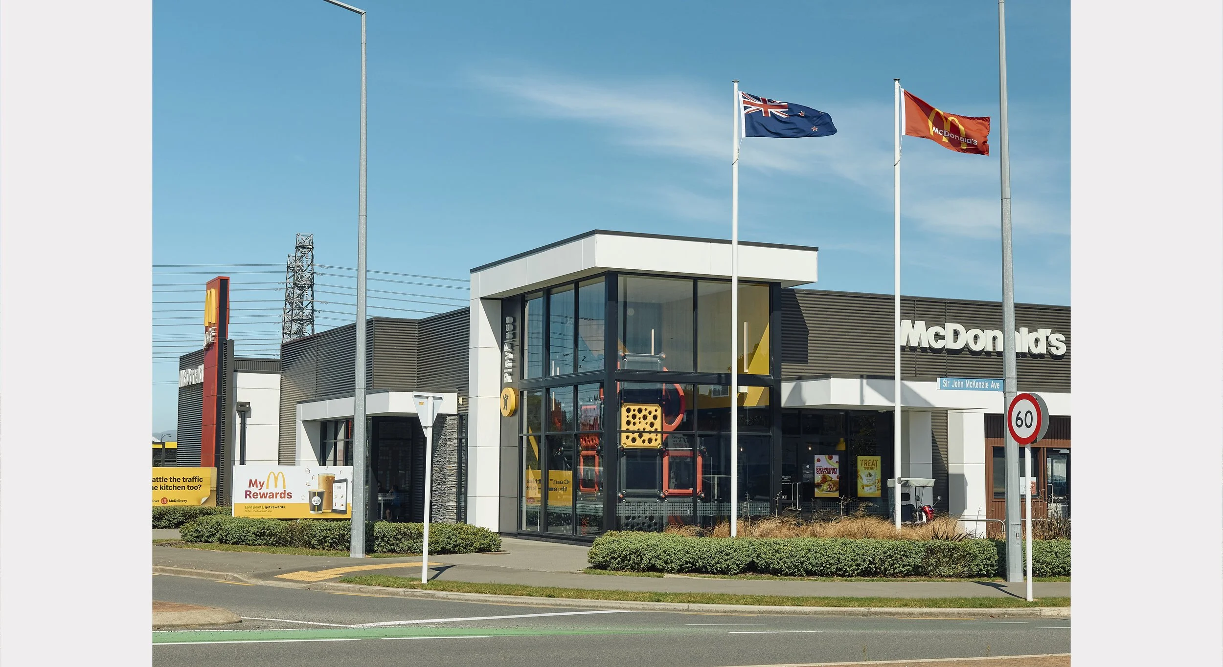 A McDonald's fast-food restaurant with three flags flying on poles, including a New Zealand flag, a McDonald's flag, and a red flag. The building has modern architecture with large glass windows and a sign that reads "McDonald's." There are cars parked outside, and the street name "Sir John McKenzie Ave" with a speed limit sign of 60 km/h.
