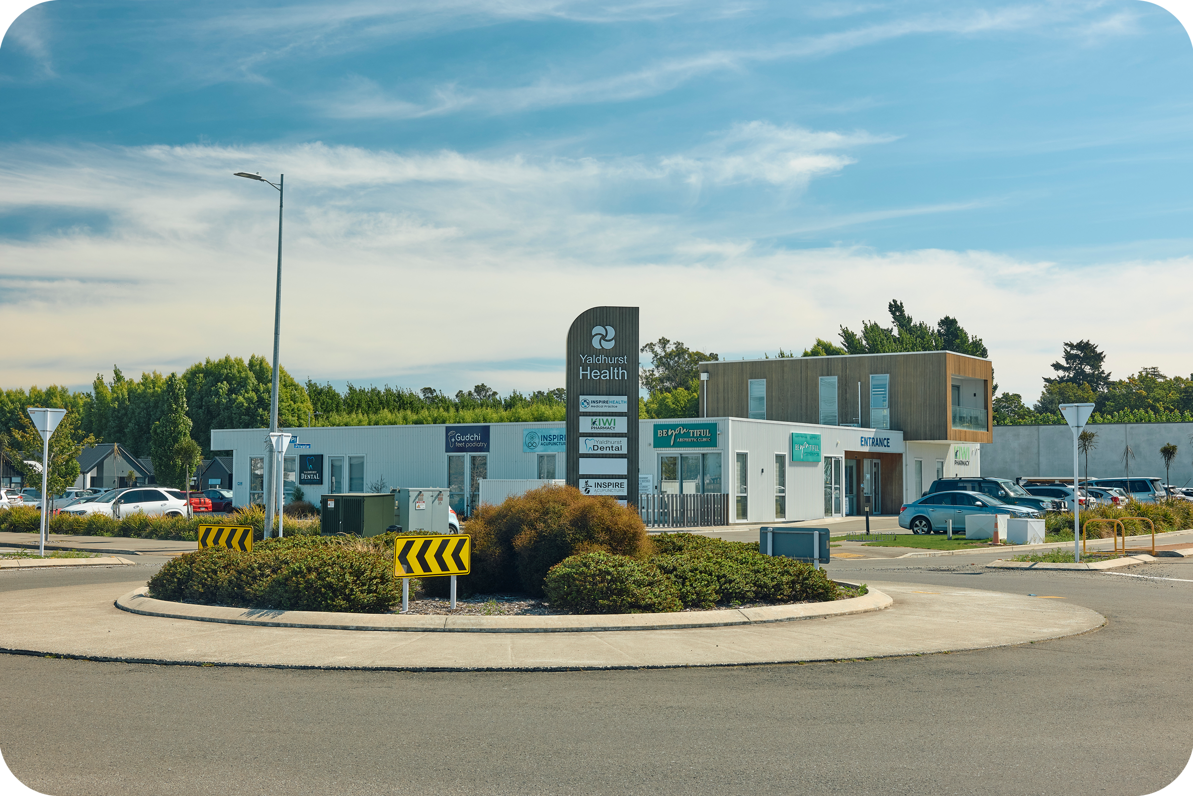 A parking lot in front of a building with signs indicating it is Yaldhurst Health. The building has a modern design with wood and glass elements. Multiple cars are parked in the lot, and there are trees and greenery around the area. The sky is blue w