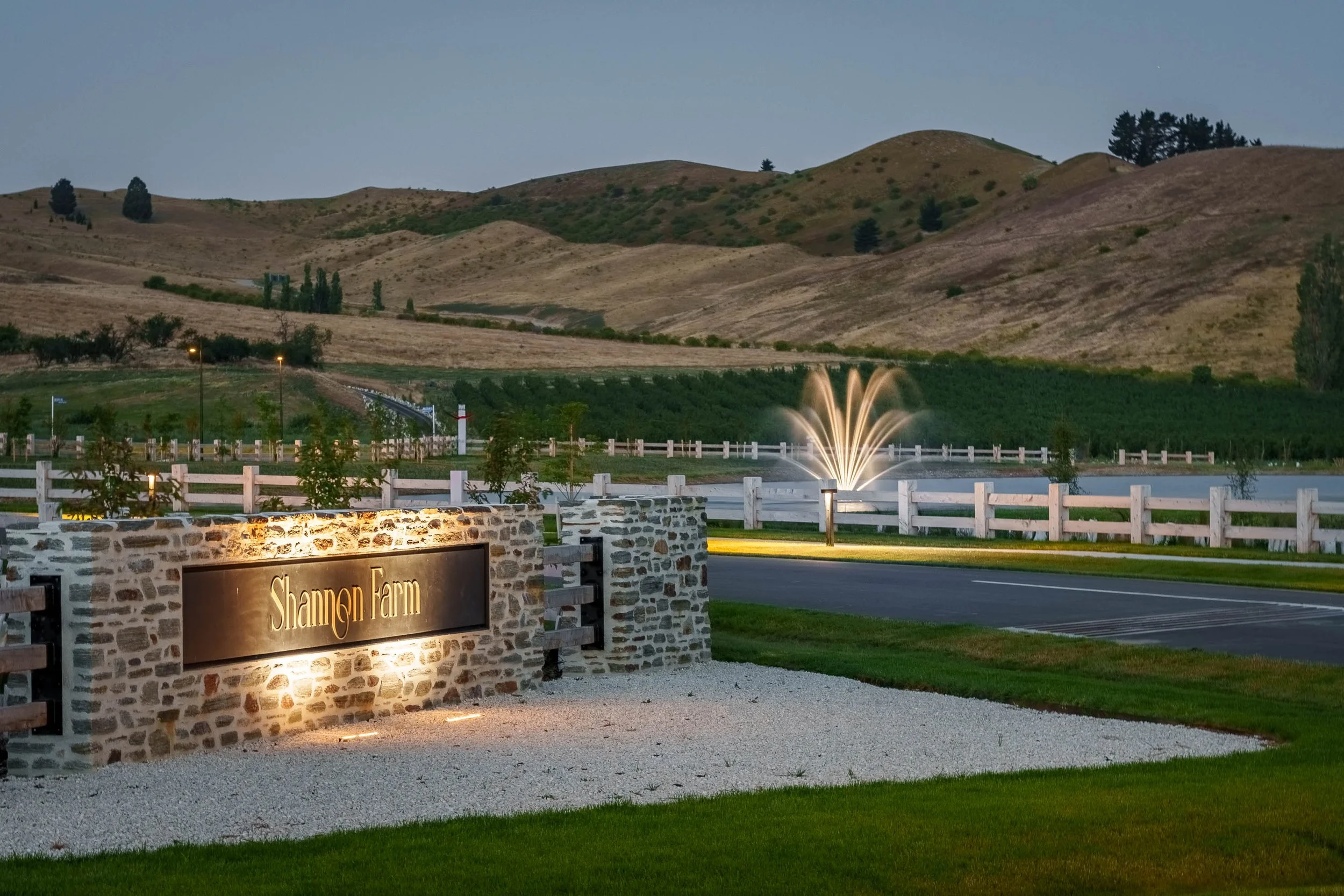 Entrance sign for Shannon Farm with a stone wall and lighting, with a fountain in a pond, farmland, and rolling hills in the background during dusk.