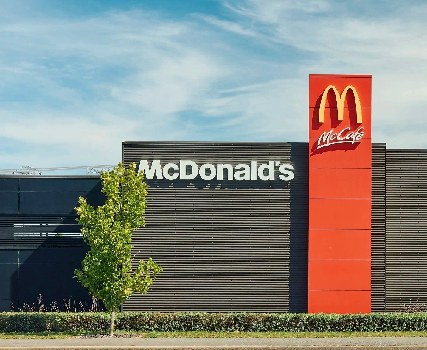 Exterior of a McDonald's restaurant with a sign displaying the McDonald's logo and a separate sign with the McCafé logo, a black and orange building, a small green tree, and a clear blue sky.