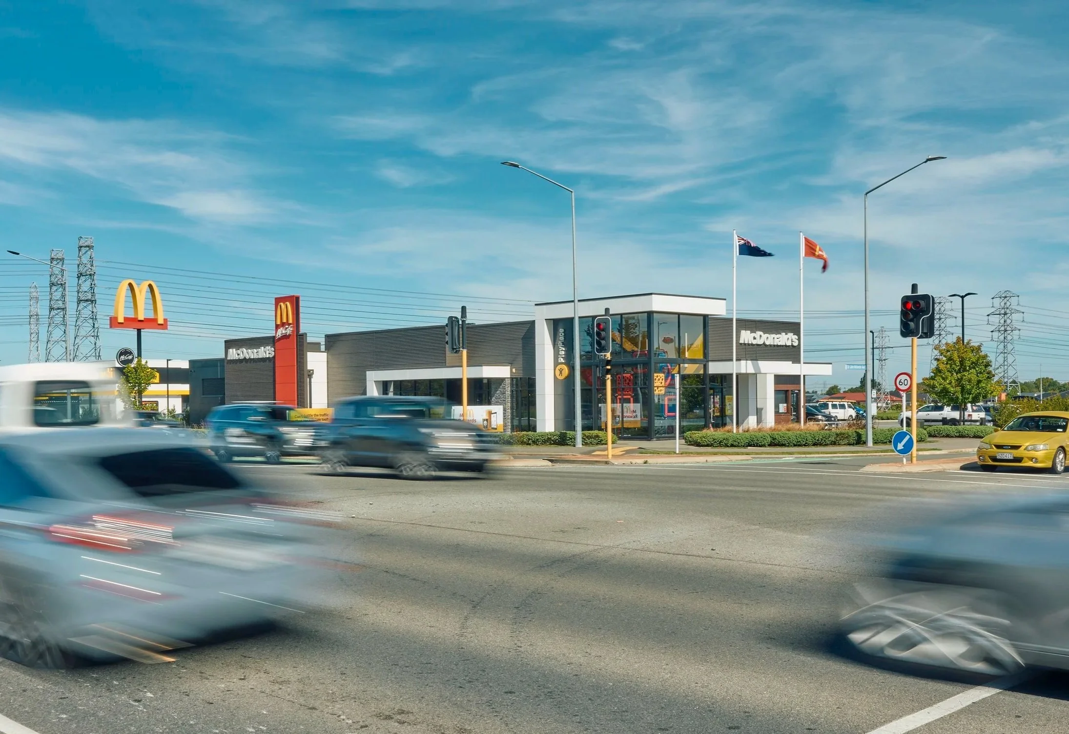 View of a McDonald's fast food restaurant with a drive-thru, traffic lights, and cars driving by on a busy street under a blue sky with some clouds.