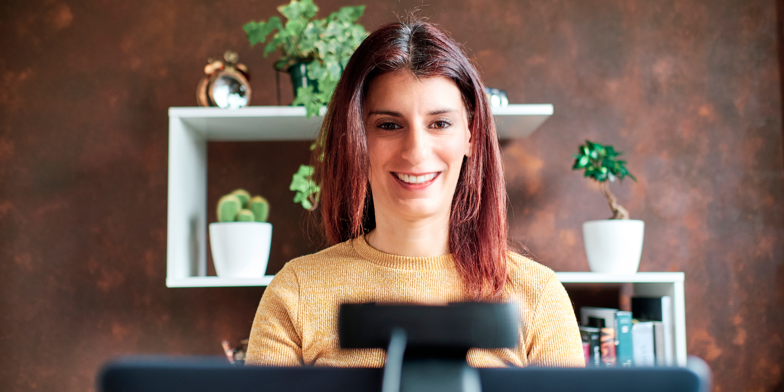Woman sitting in front of a computer with the camera on. Shelving with books and plants is behind her.