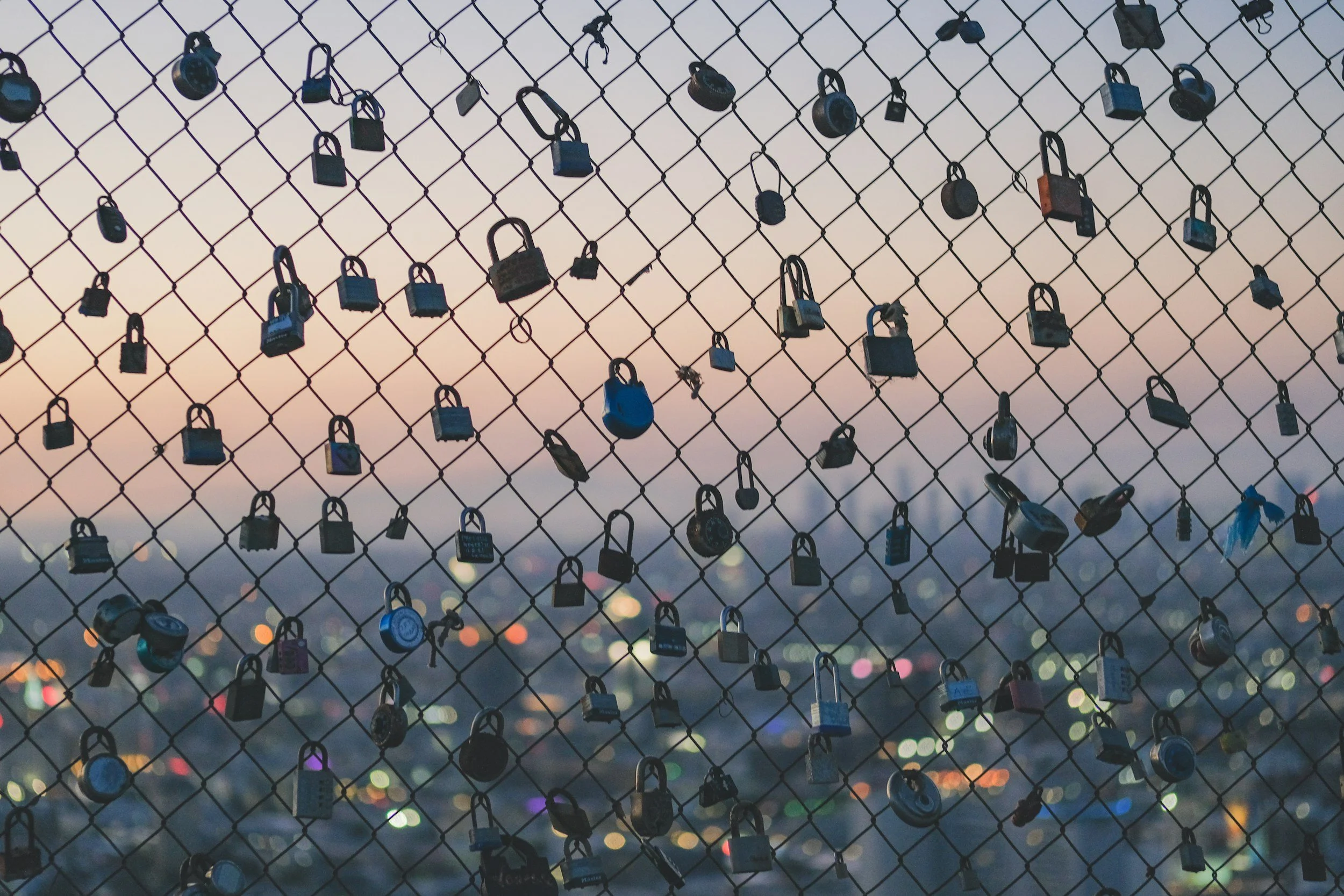 A chain-link fence with numerous padlocks attached, set against a blurred cityscape and sunset sky in the background, representing the security of personal data at Gembridge.