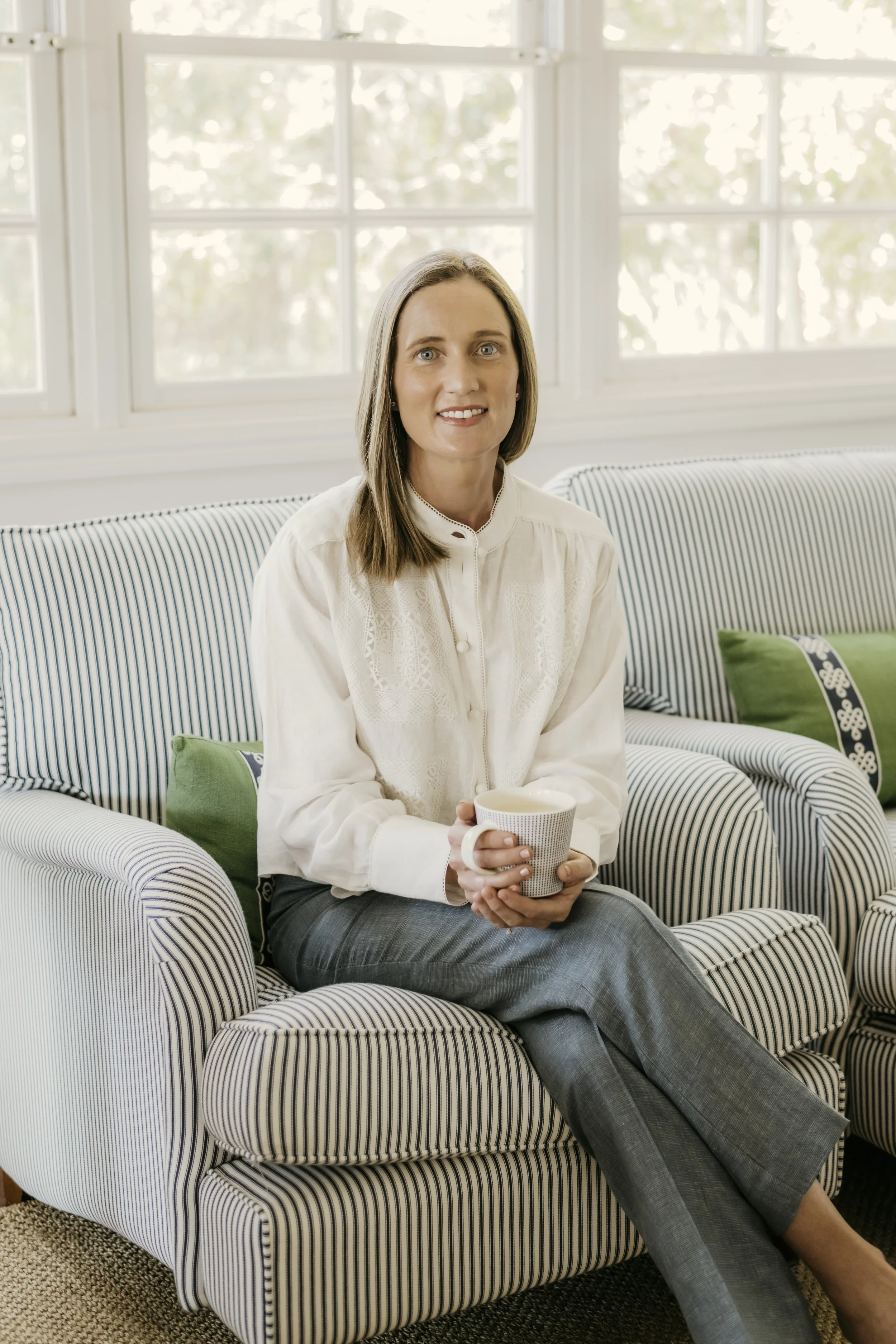 A woman with shoulder-length brown hair and blue eyes sitting on a striped couch, holding a mug, in front of a large window with trees outside.