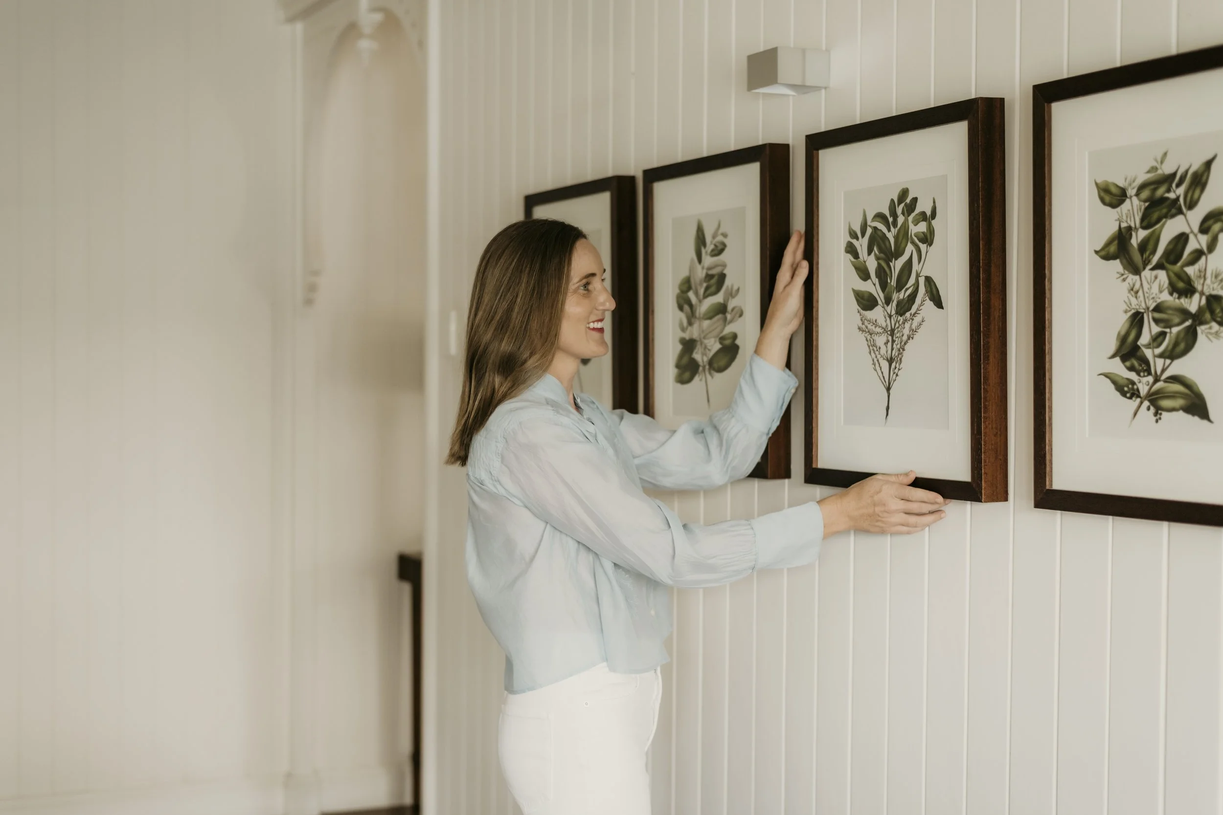 A woman with brown hair and a light blue blouse arranging framed botanical art prints on a white wall.