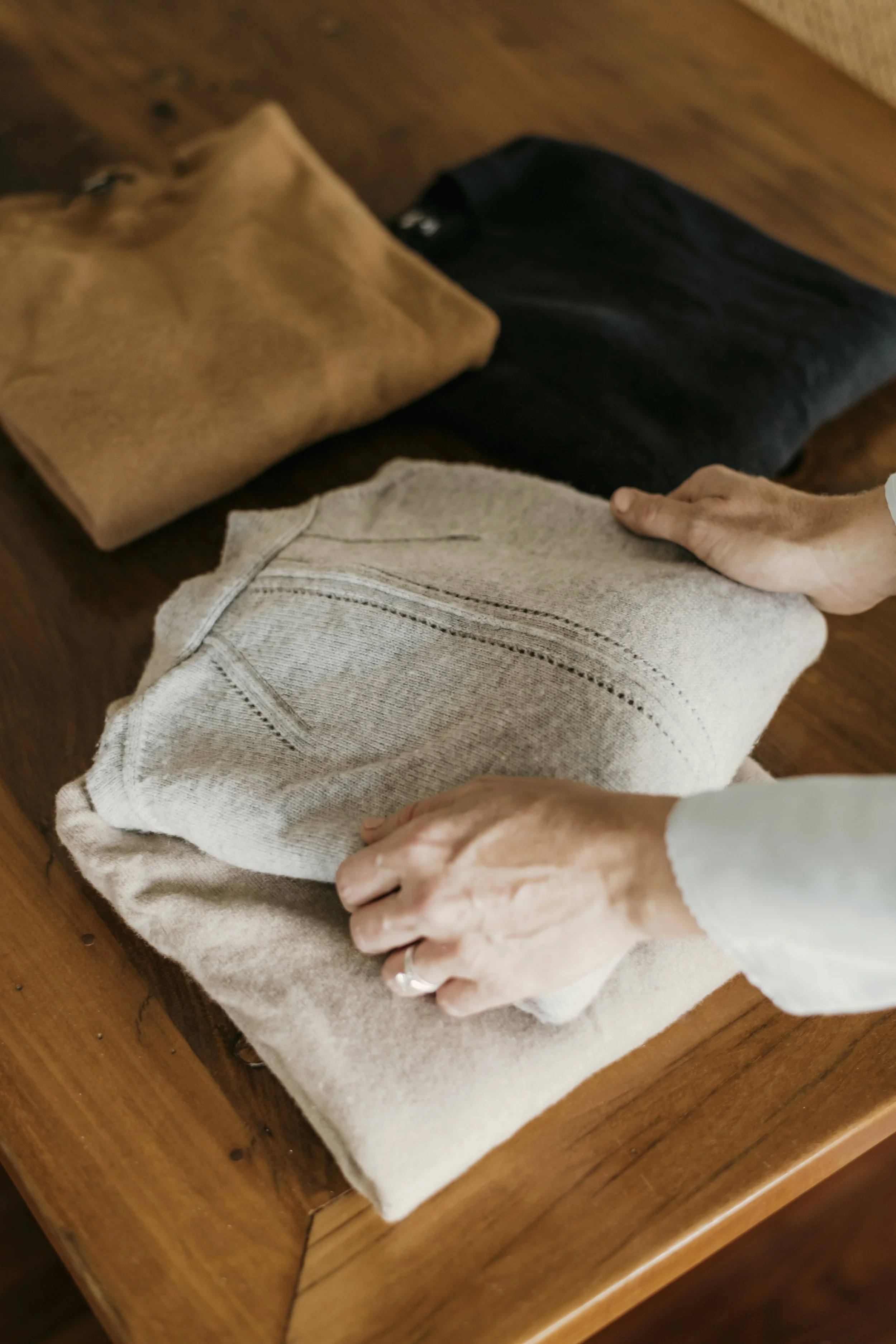 Person folding a beige cloth on a wooden table with other folded clothes nearby.