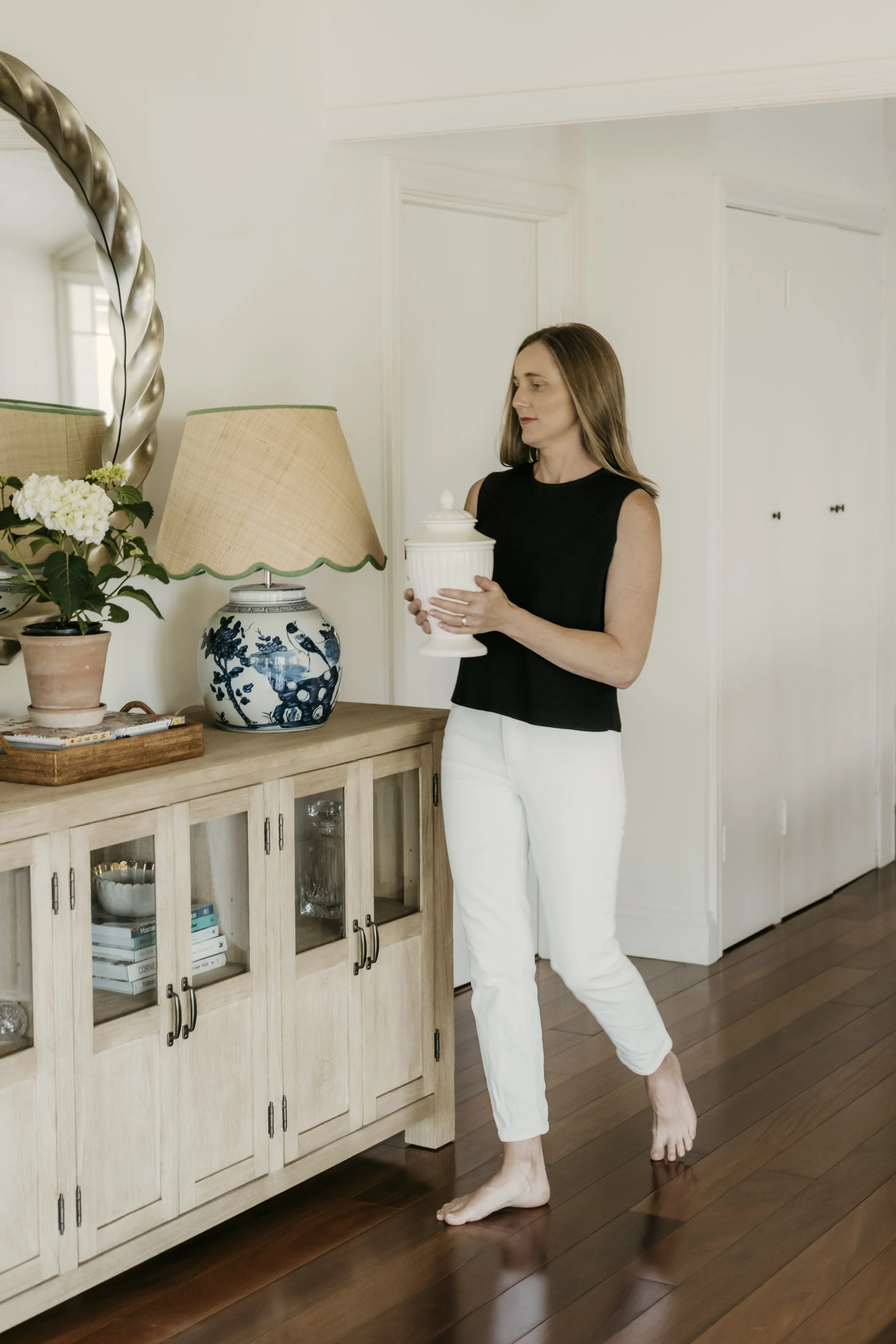 Woman standing barefoot in front of a wooden sideboard, holding a white ceramic container with a lid, dressed in a black sleeveless top and white pants, in a bright, minimalistic home with white walls and wooden floors.