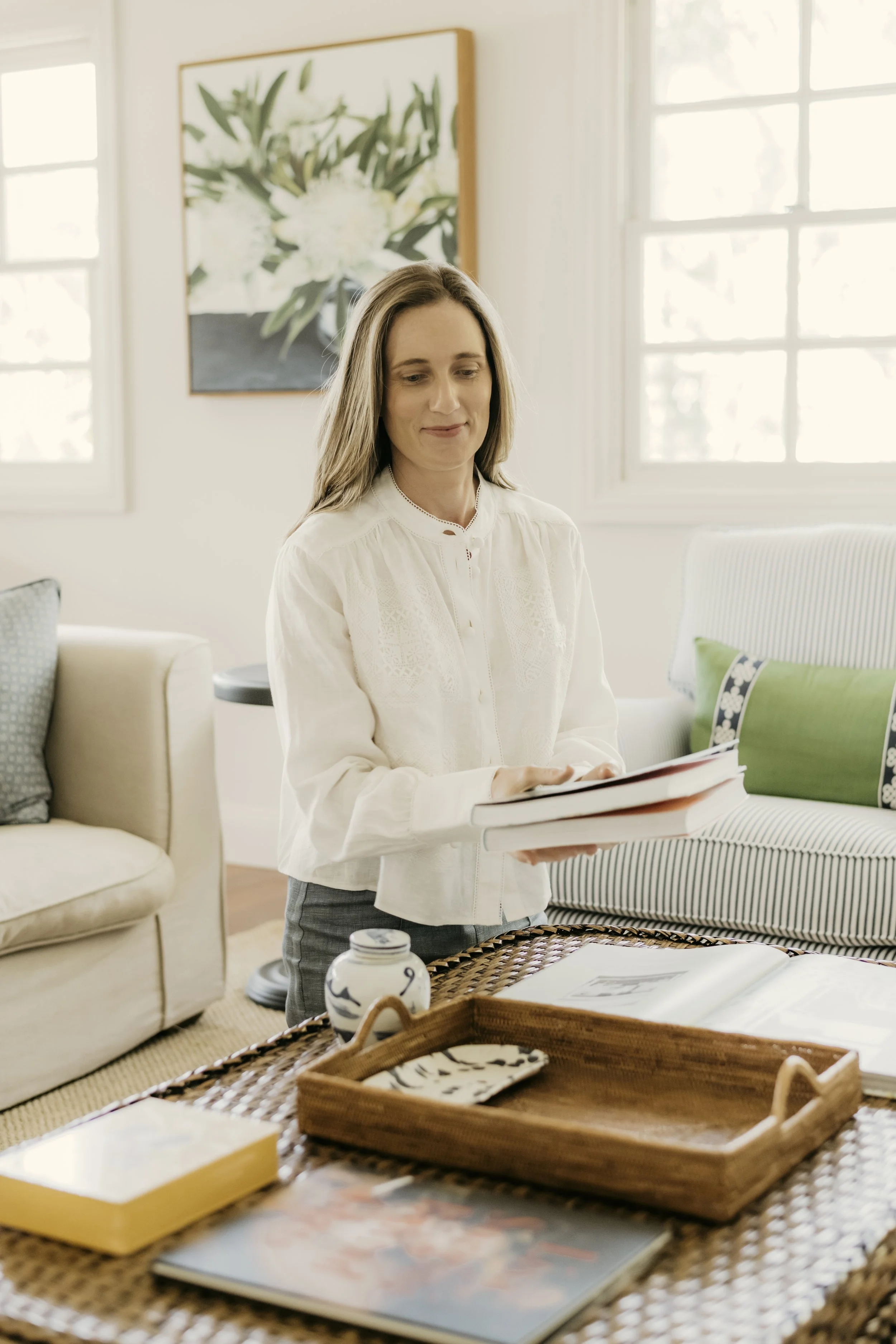 A woman with long light brown hair wearing a white blouse holding a book in a bright living room with beige furniture, plants, and artwork on the wall.