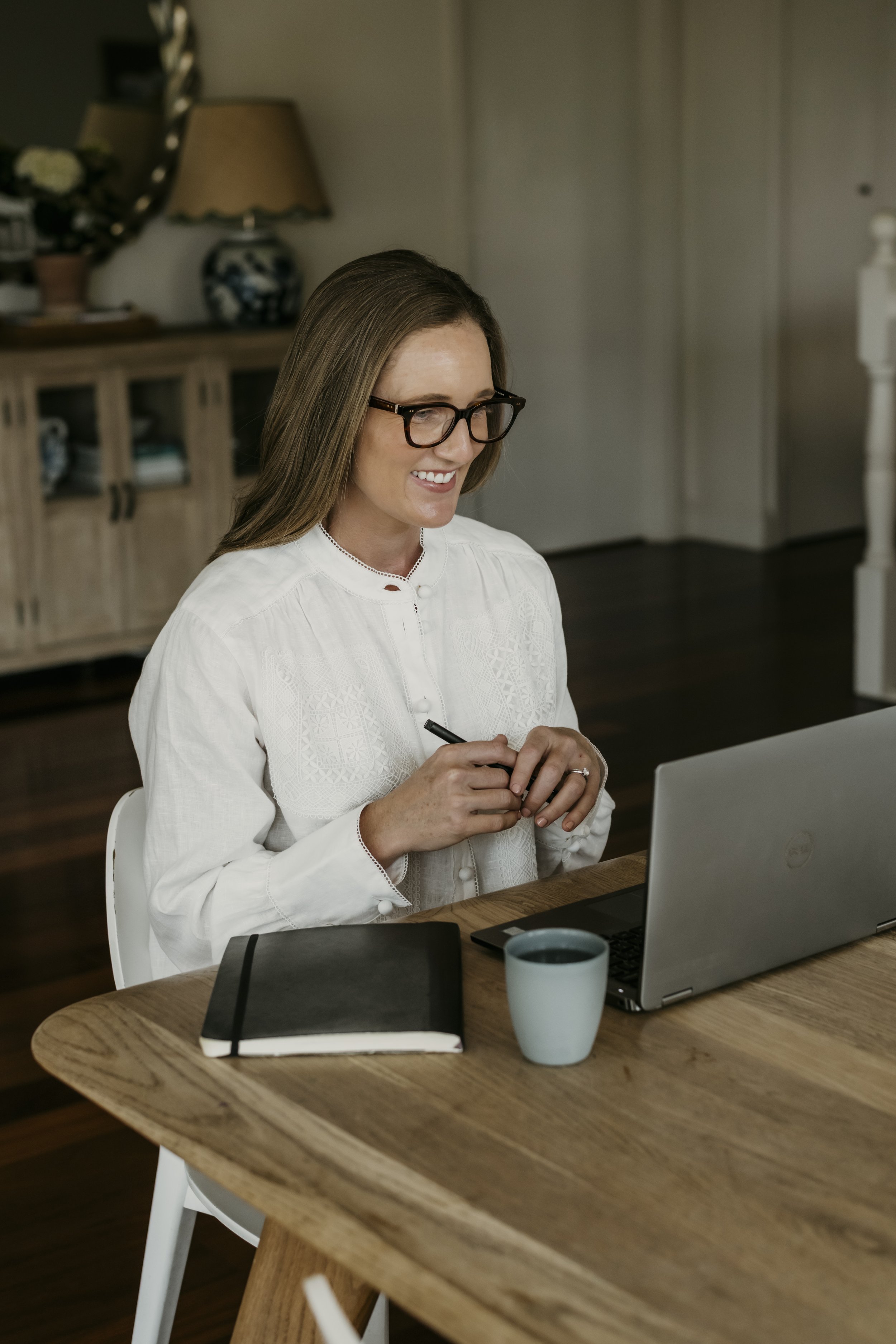 A woman sitting at a wooden table with a laptop, a notebook, a pen, a mug, and a tablet, working and smiling.