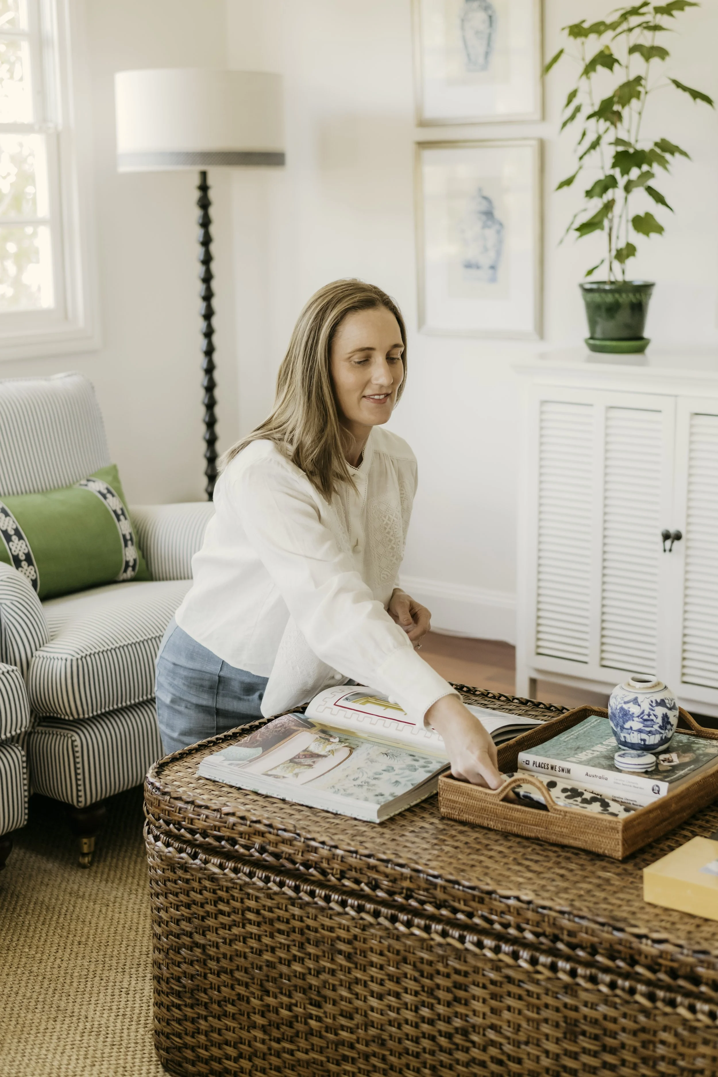 A woman with long light brown hair, wearing a white blouse, is reaching into a collection of magazines and books on a wicker coffee table in a bright, airy living room with natural light. The room has a white striped sofa with green and black pillows, a white radiator cover, a tall potted plant, and framed artwork on the wall.