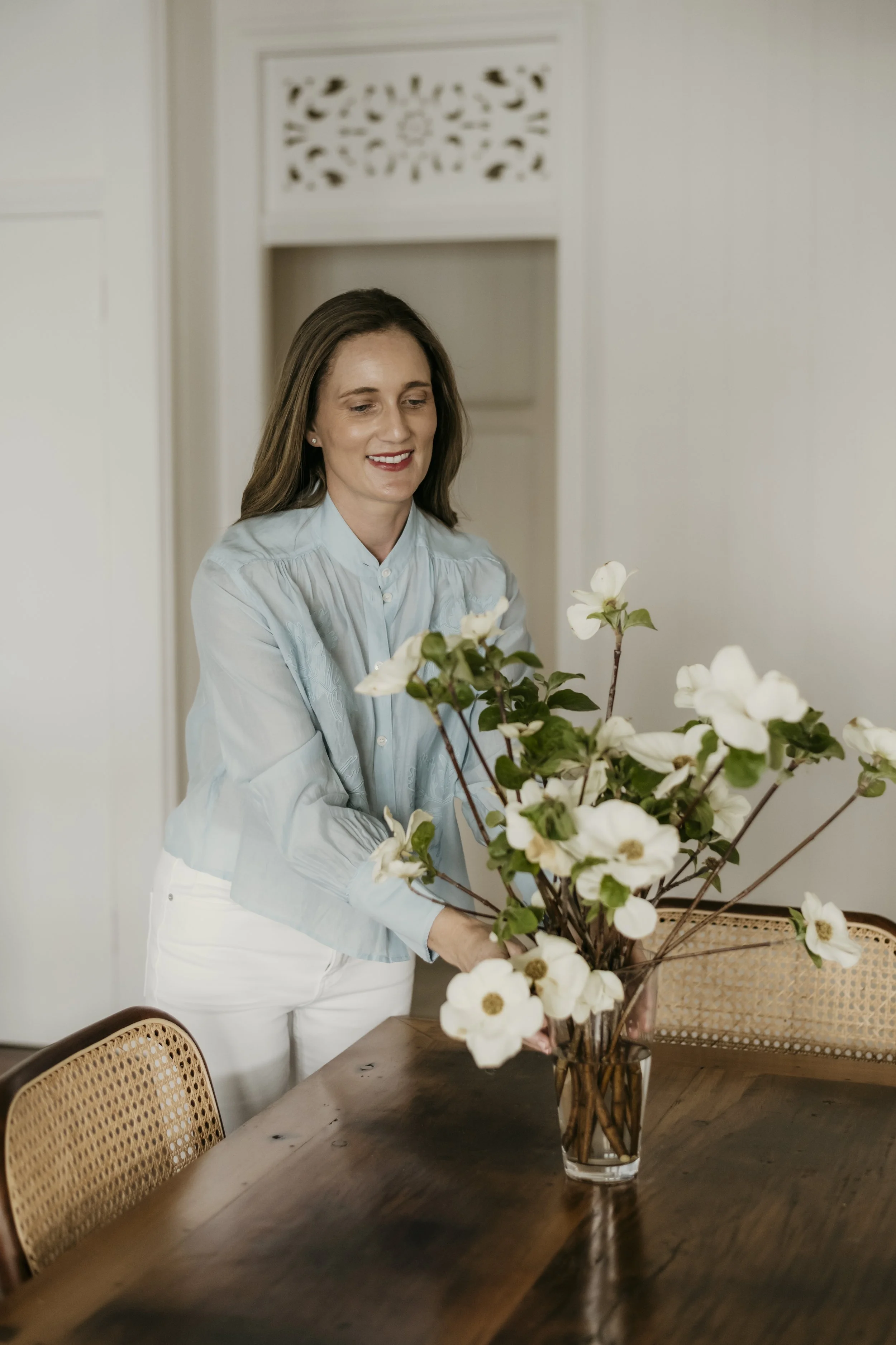 A woman with long brown hair, wearing a light blue button-up shirt and white pants, arranging white flowers in a tall glass vase on a wooden dining table in a bright, modern room.