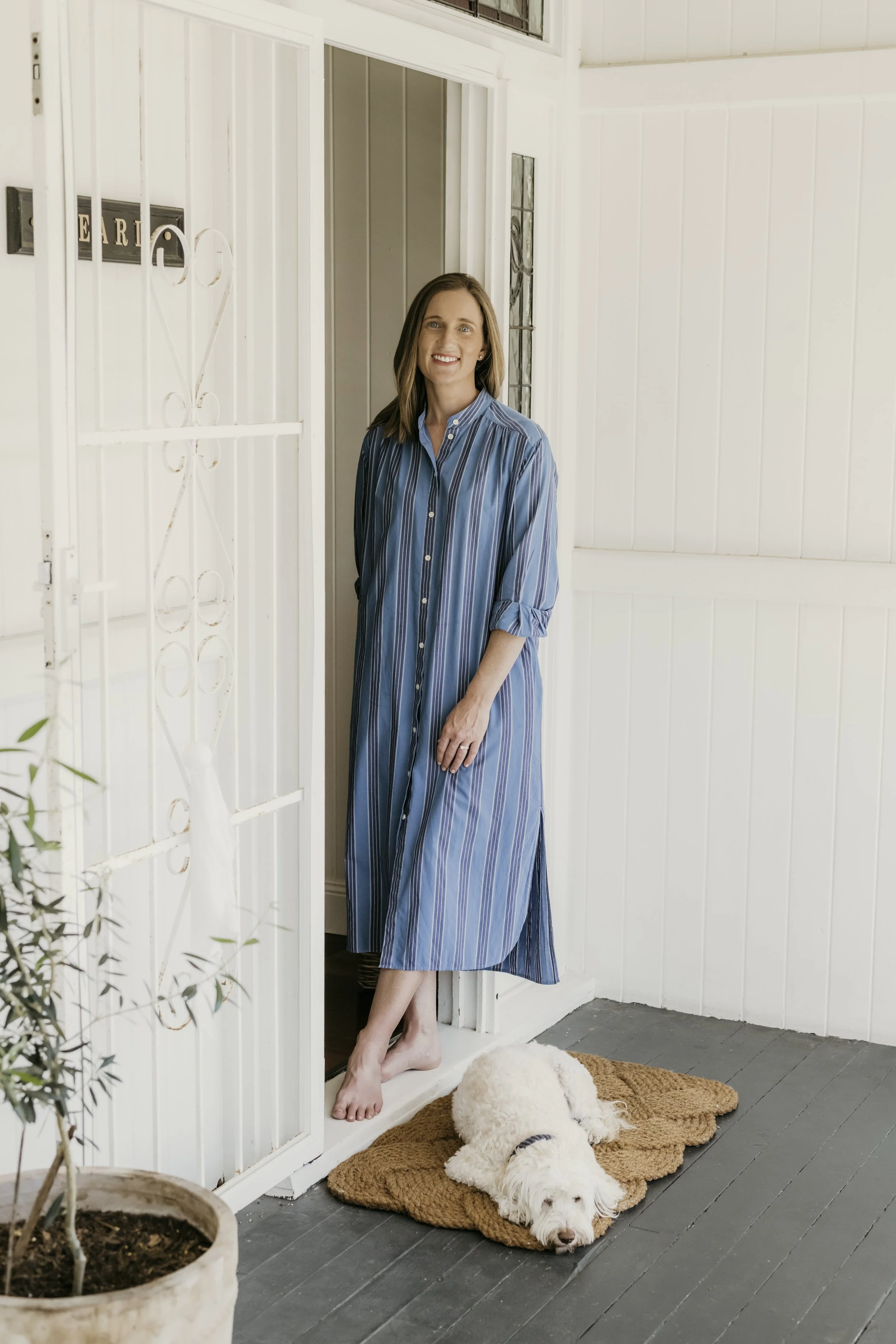 A woman in a blue striped dress standing on a porch with a dog lying on a brown rug in front of her. The porch has white walls and a gray floor. A potted plant is visible in the foreground, and the dog appears to be resting peacefully.