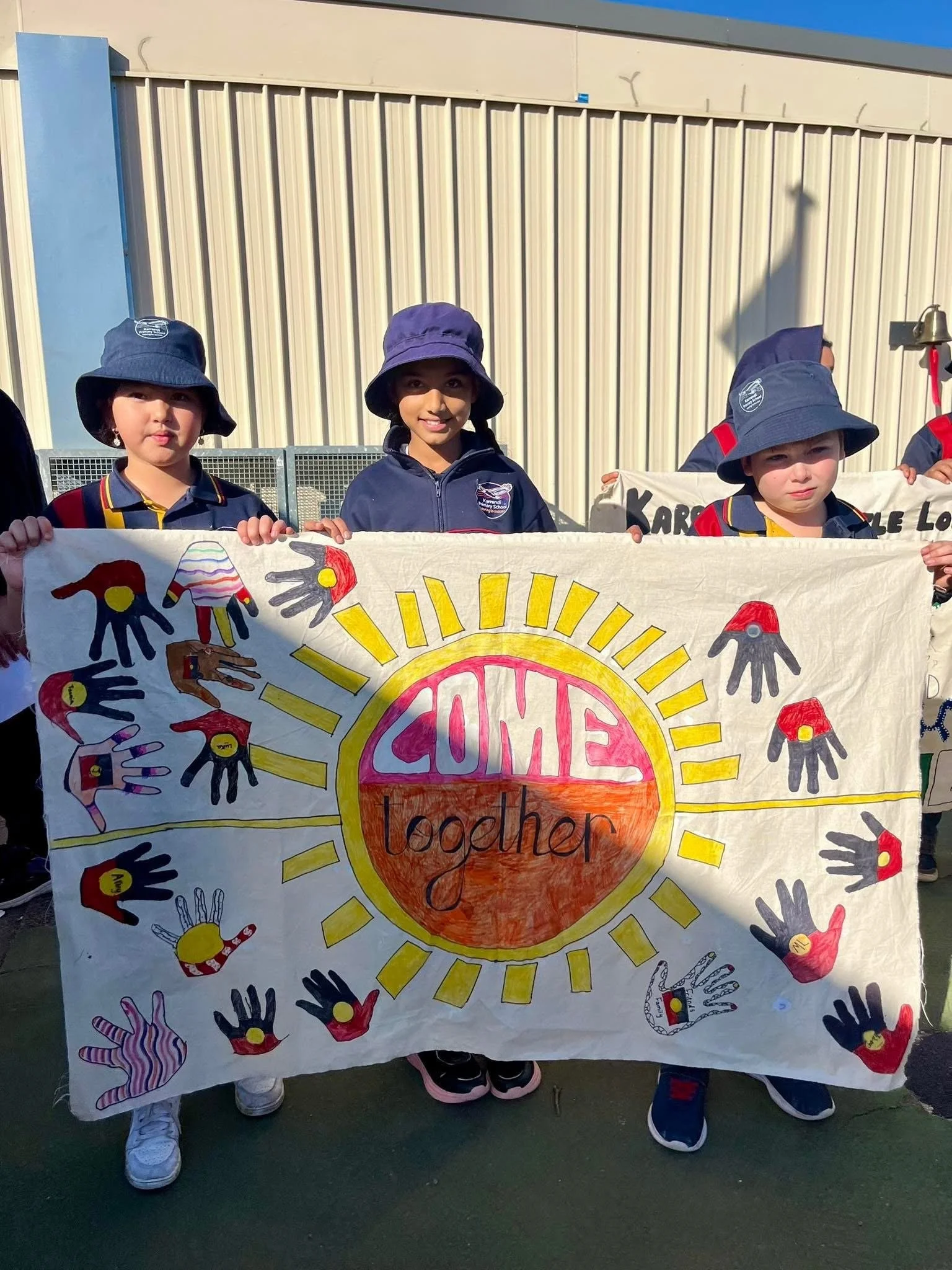 Young children holding a colorful banner with the words 'Come Together' on it, surrounded by various handprint designs.