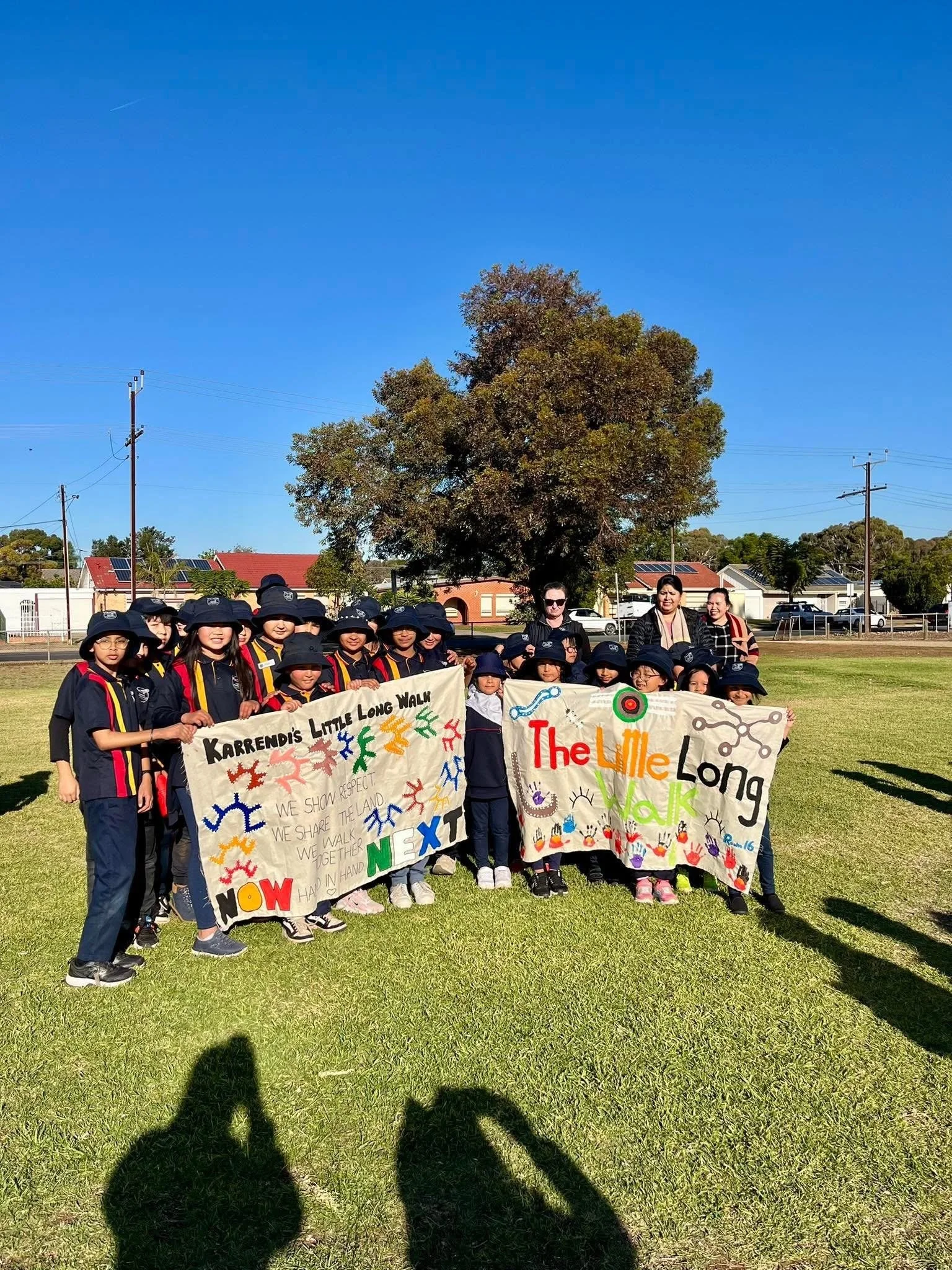 A group of children and adults holding colorful banners for a walk event outdoors on a sunny day, with trees and houses in the background.