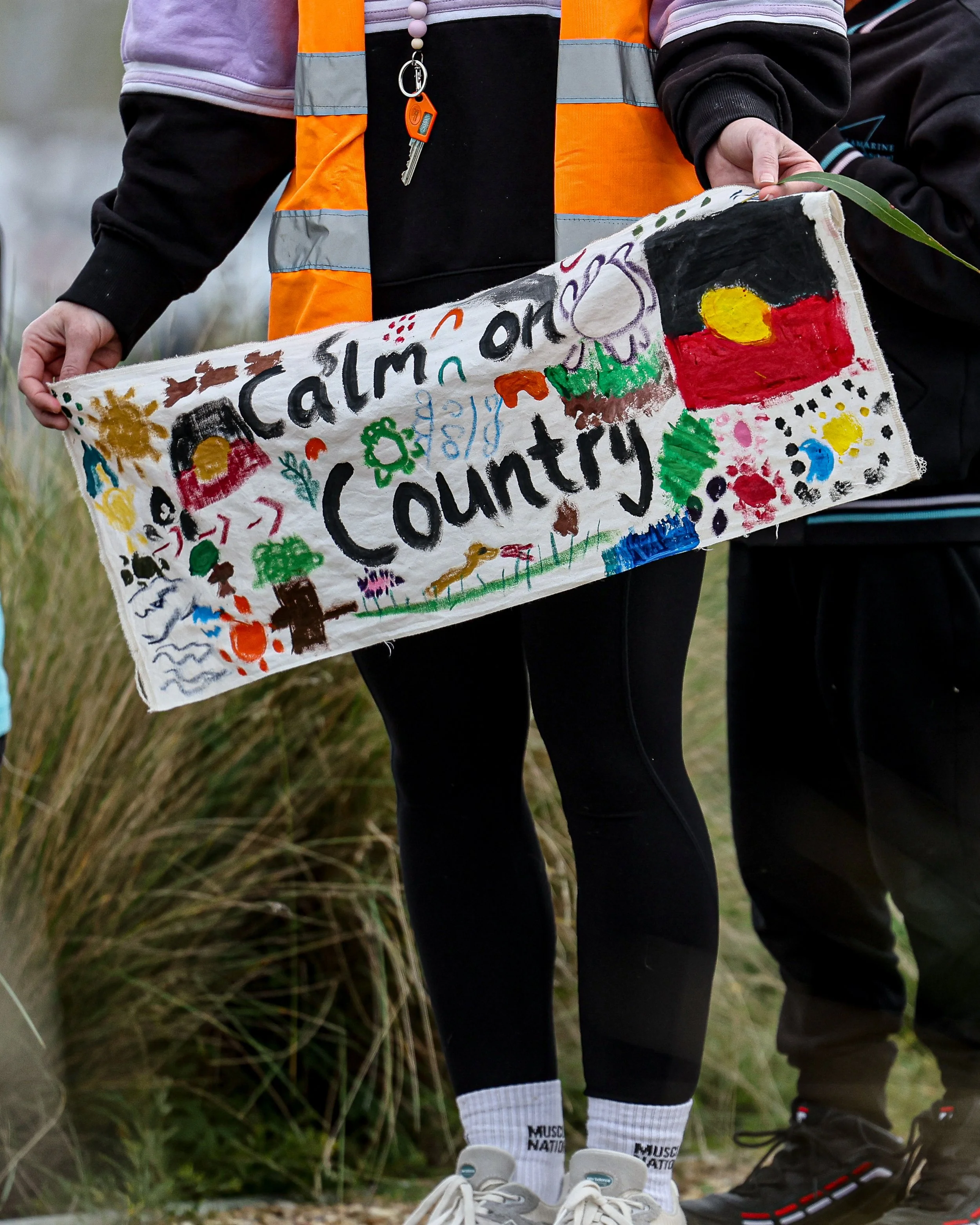 Person holding a colorful, hand-painted fabric banner with the words 'calm on country' and various painted symbols and images, dressed in outdoor athletic gear.