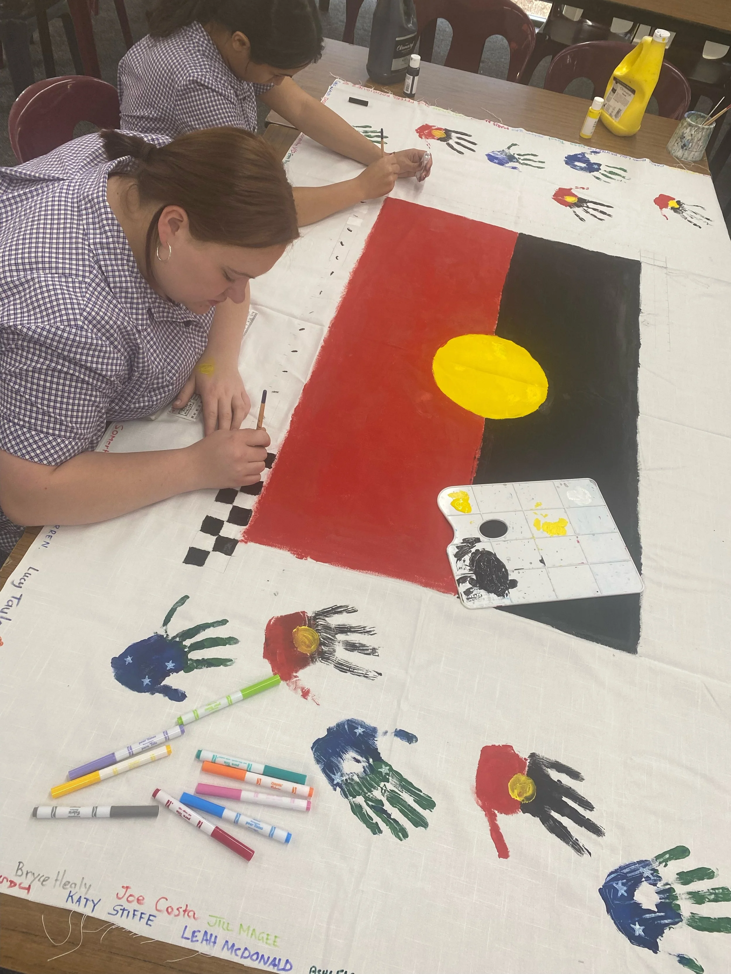 Two girls in checkered shirts are painting a large flag on a white cloth. The flag has red, black, and yellow sections with a large yellow circle in the middle. There are various colored handprints on the cloth, as well as paint supplies and markers 