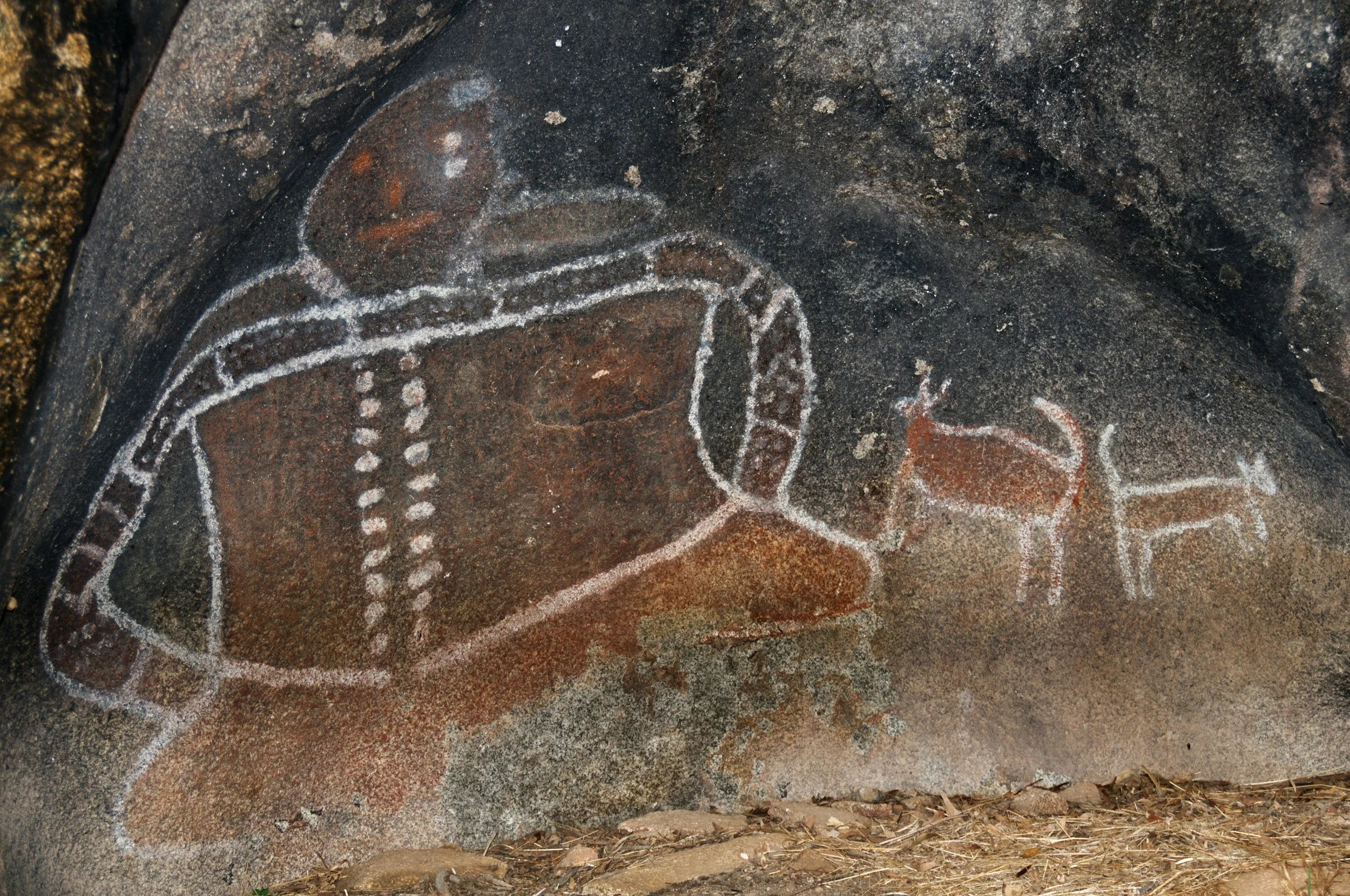 Ancient rock art depicting a human figure with elongated limbs, detailed with white chalk lines, on a rugged dark stone surface with surrounding natural earthy tones.