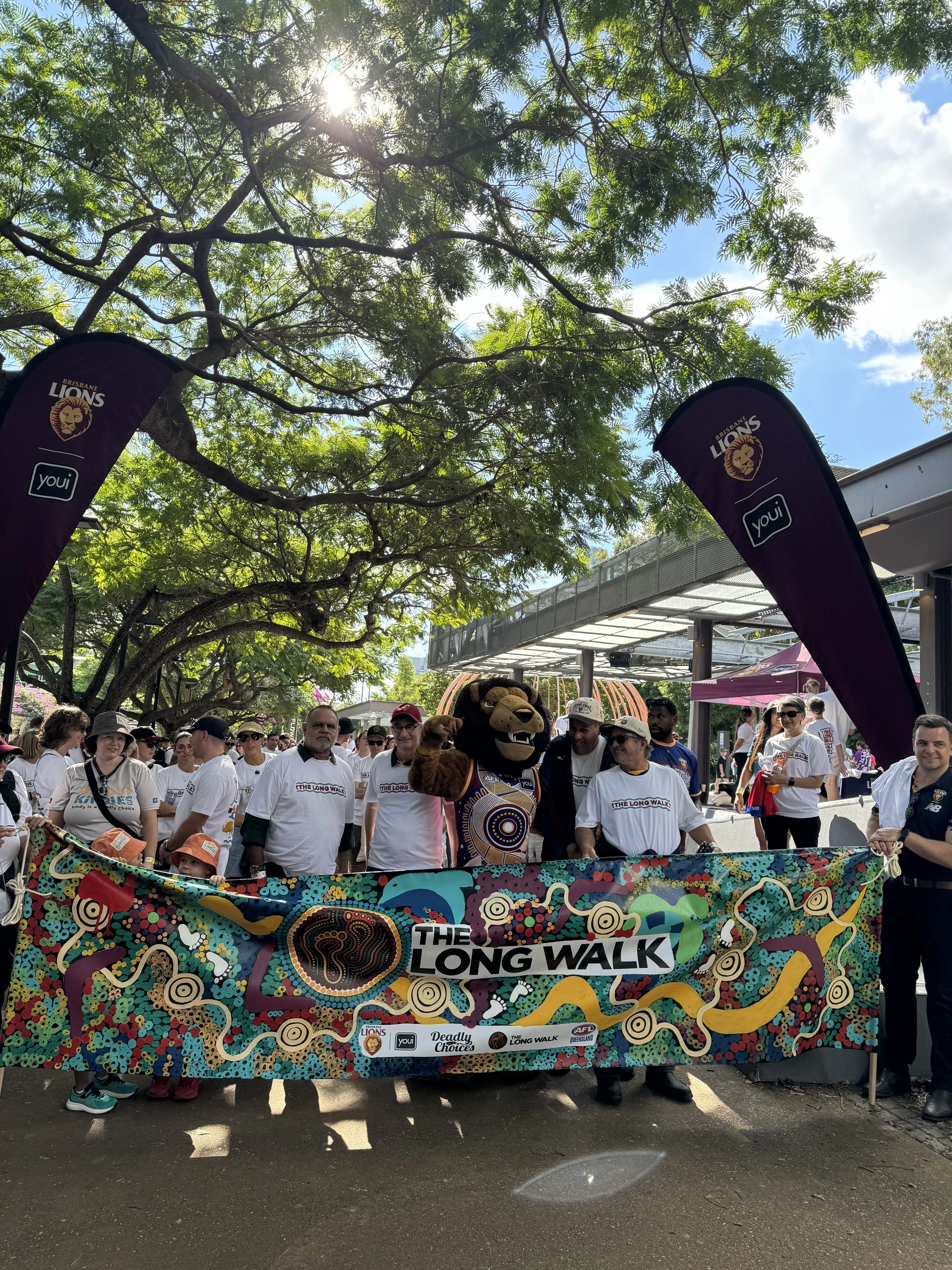 A large group of people participating in a parade or event, holding a colorful banner with the words "THE LONG WALK" on it. Some people are wearing white T-shirts, and a mascot or costumed character resembling a lion is present in the crowd. There ar