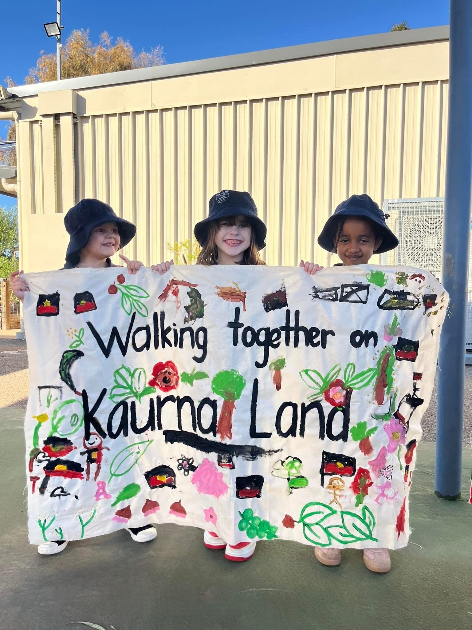 Three children dressed in black hats and pajamas holding a large, colorful banner with drawings of strawberries, leaves, and flowers, and the words "Walking Together on Kauru Land" written on it, outdoors during daytime.