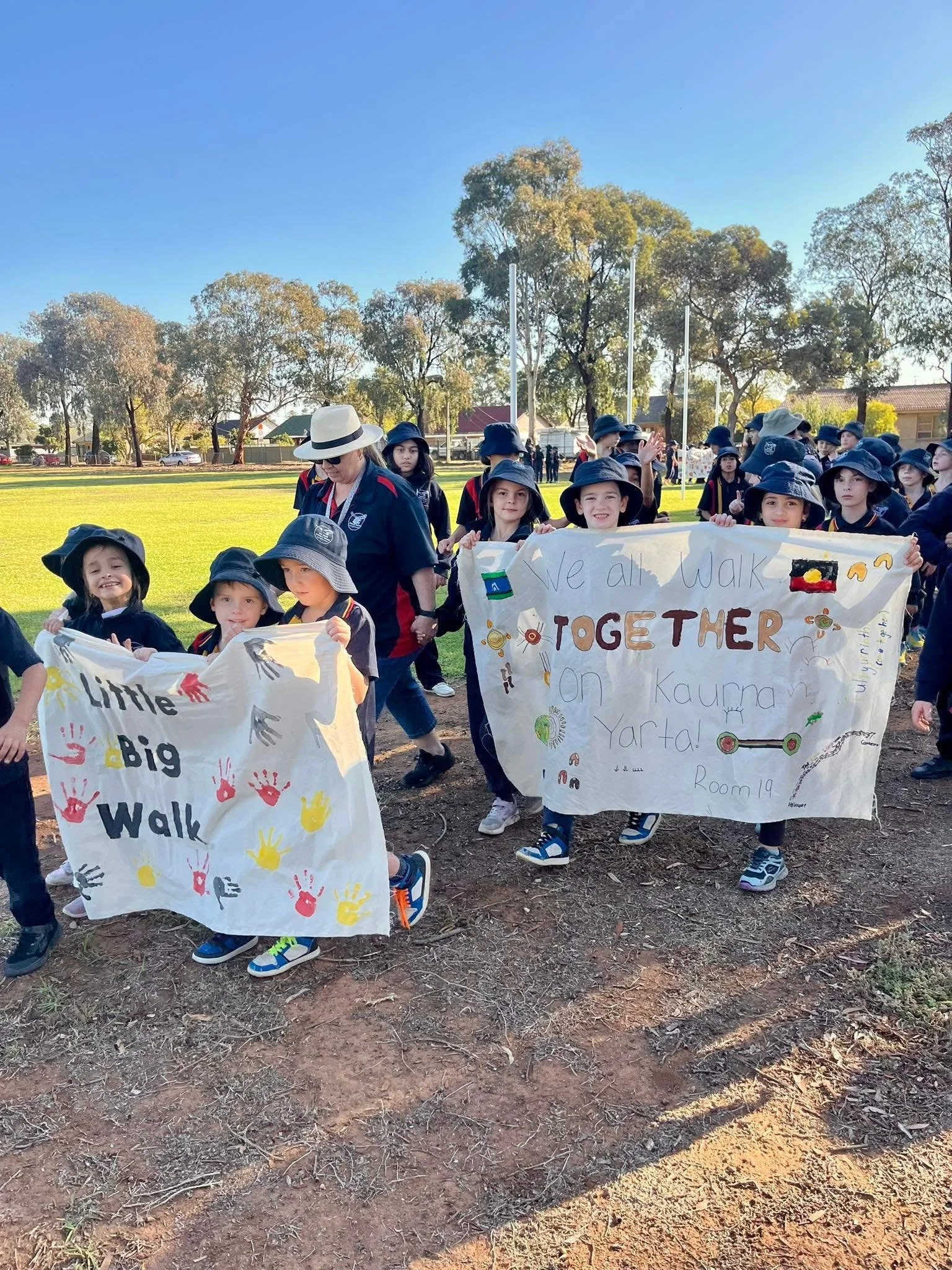 A group of children and a woman holding handmade signs and participating in an outdoor walk event on a grassy field with trees, houses, and a clear blue sky in the background.