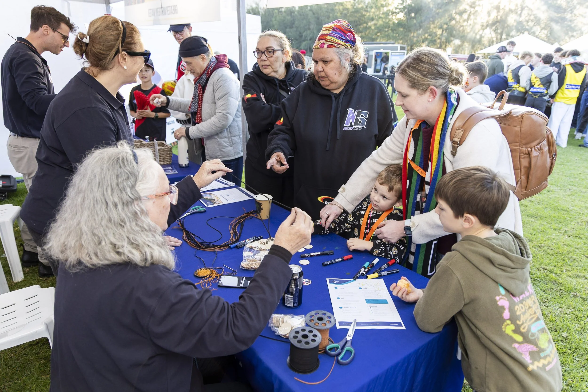 People gathered around a table at an outdoor event, engaging in a hands-on activity involving wires and electronic components.