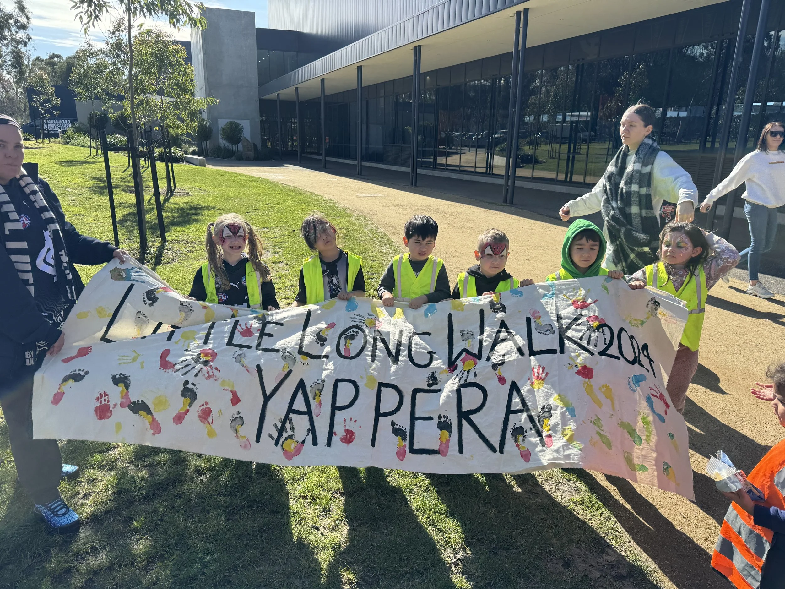 Children with face paint holding a banner at a protest or demonstration outside a modern building, with trees and grass in the background.