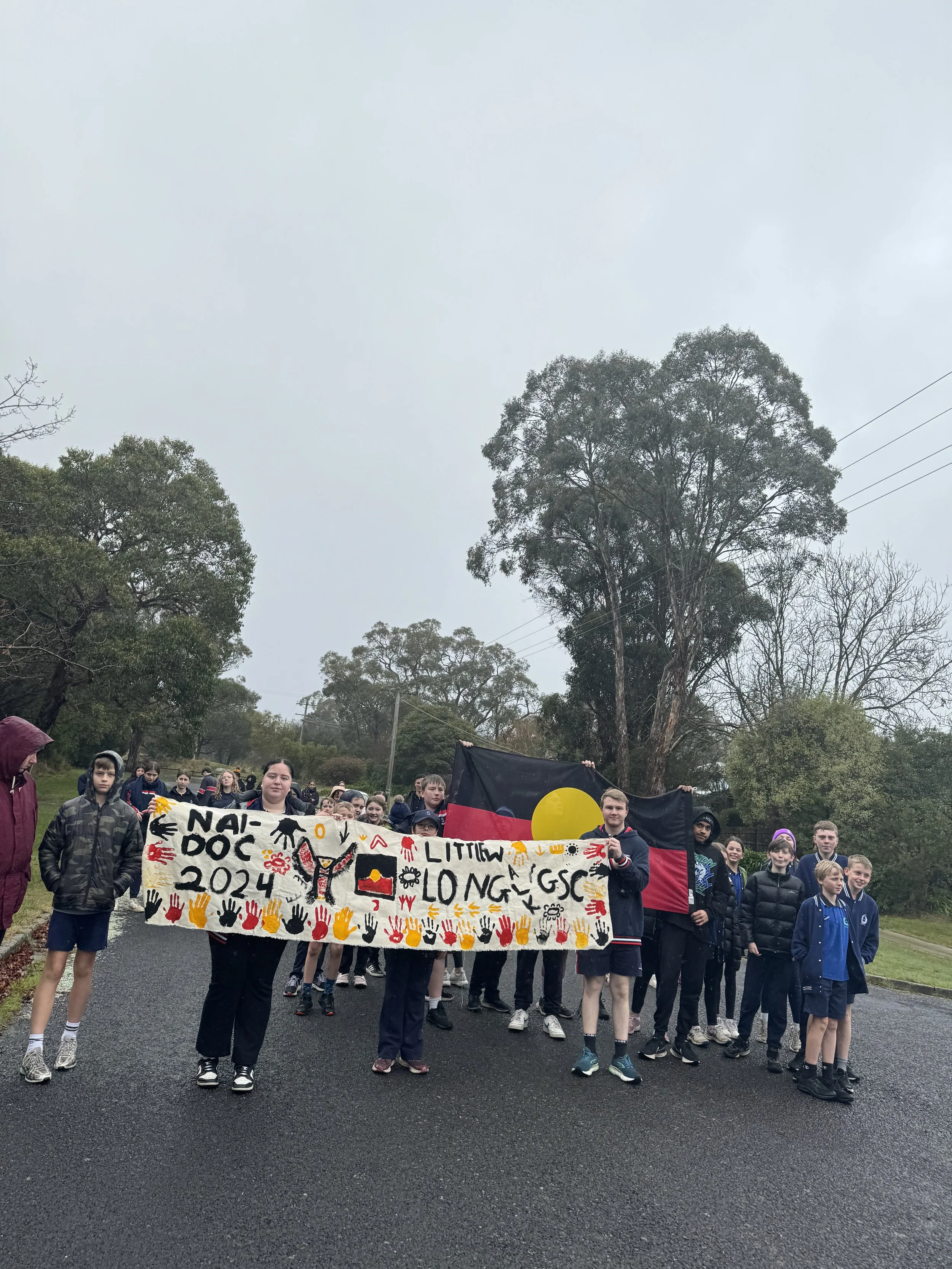 Group of children and teenagers participating in a protest or march outdoors. They are holding a colorful banner with handprints, reaching from 2024, and various symbols. Some are holding an Aboriginal flag. The sky is overcast, and there are trees a