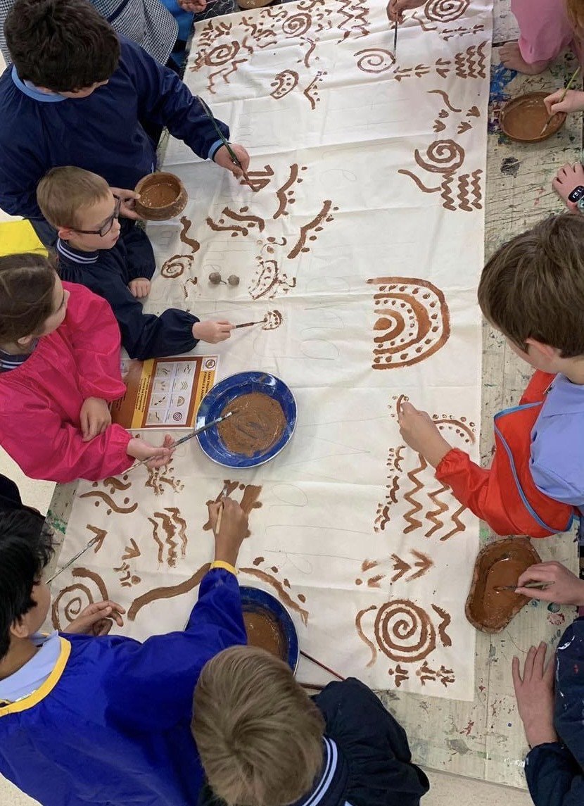 Children painting tribal or indigenous-style symbols and designs with brown paint on a large white cloth.