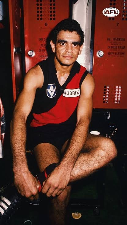 A male athlete sitting in a locker room, wearing a black and red sports uniform with a name tag 'NUBRIK.' He has dark, curly hair and is looking at the camera. There are red lockers in the background with white labels and letters.