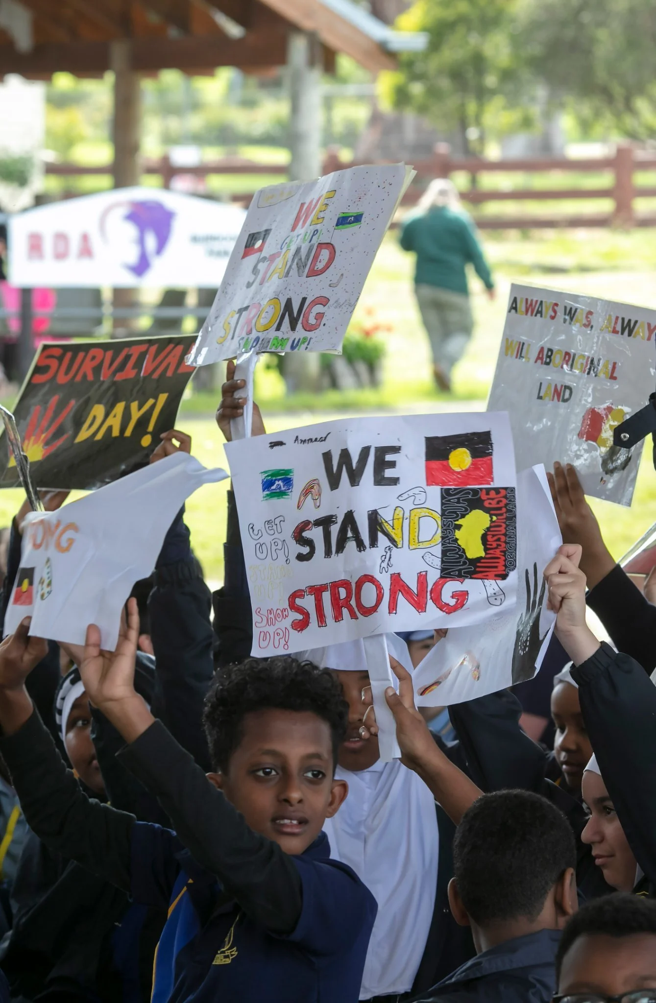 Children and young people participating in a protest or rally, holding handmade signs with messages such as "We Stand Strong", "Survival Day!", and "Always Was Always Will Aboriginal Land". The scene is outdoors with trees and a fence in the backgrou