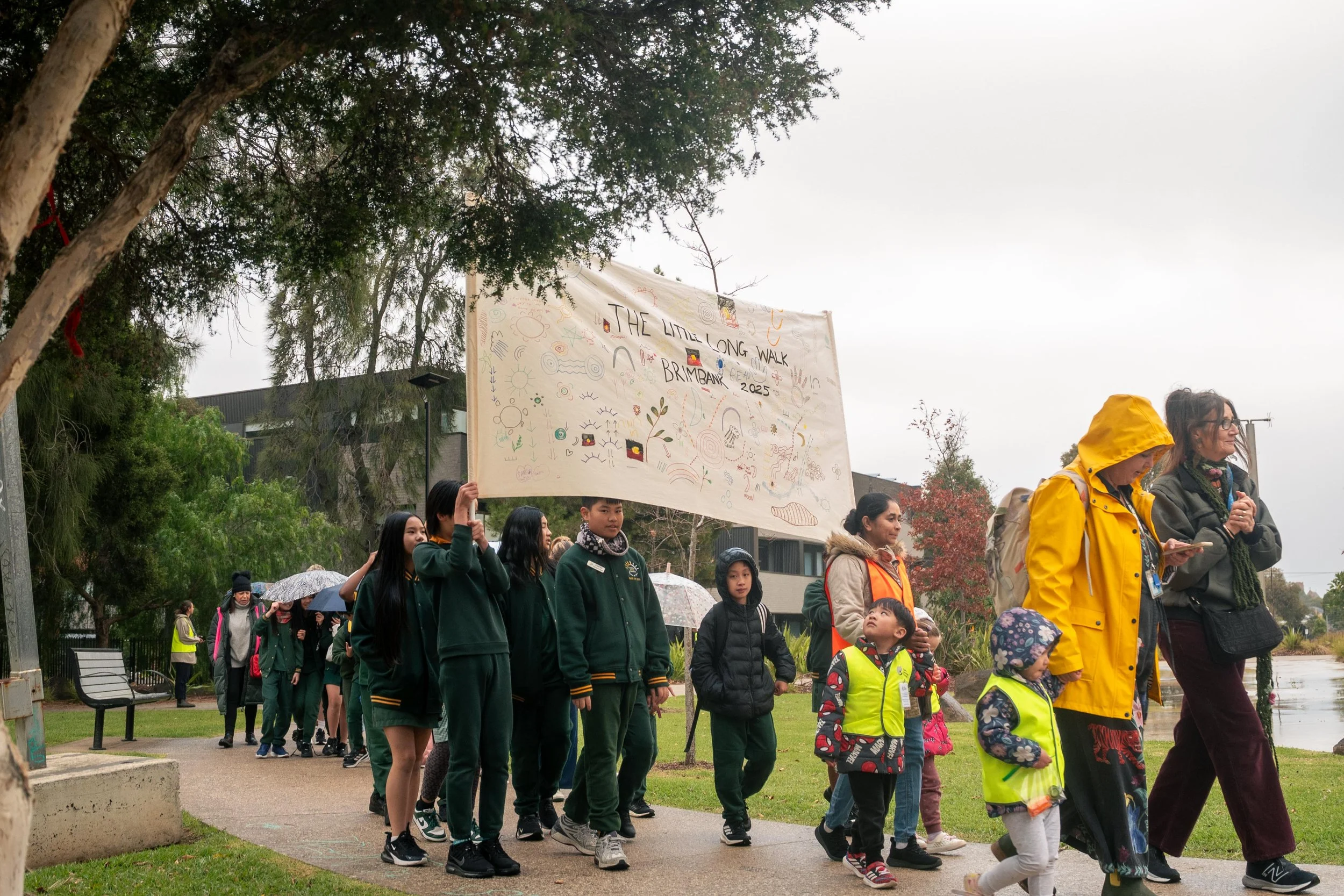 Children and adults in a park participating in a march or gathering. Some children wear bright yellow jackets and others hold umbrellas. A large banner with doodles and text, including the phrase 'The Little Long Walk,' is being held up by people in 