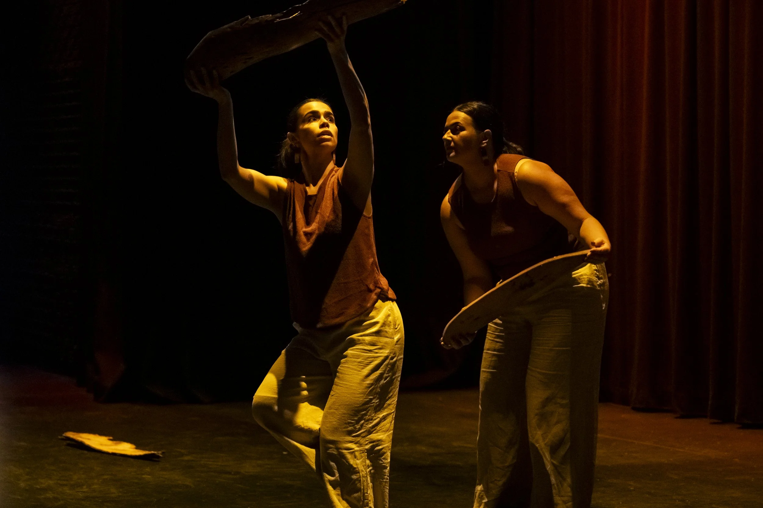Two women performing a dance on stage with dark background and warm lighting. The woman on the left holds a curved piece of wood above her head, while the woman on the right bends slightly forward holding a similar object. Both wear sleeveless costumes and yellowish pants.