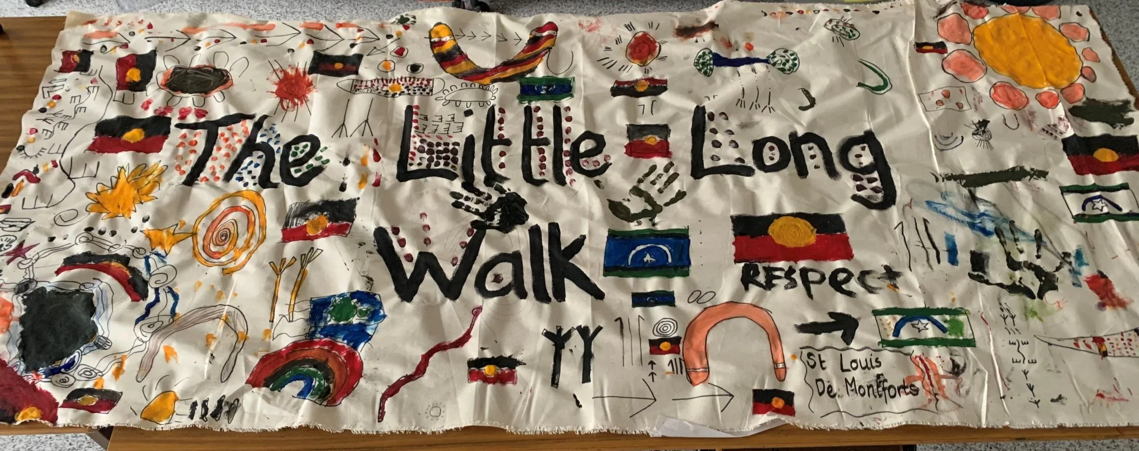 A fabric banner with colorful drawings and writings, including the words 'The Little Long Walk', flags, and various abstract designs, related to the Indigenous Peoples' Day or a similar honoring event.