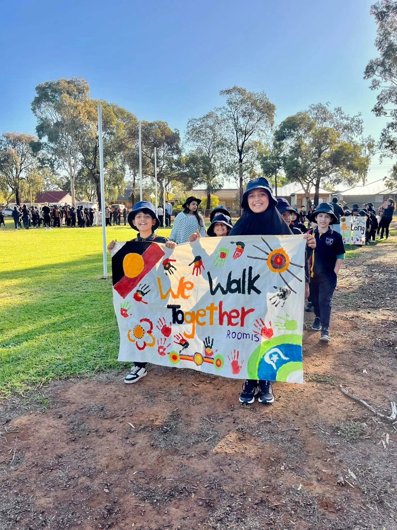 Children holding a colorful banner that says "We walk together" during an outdoor event with trees and people in the background.