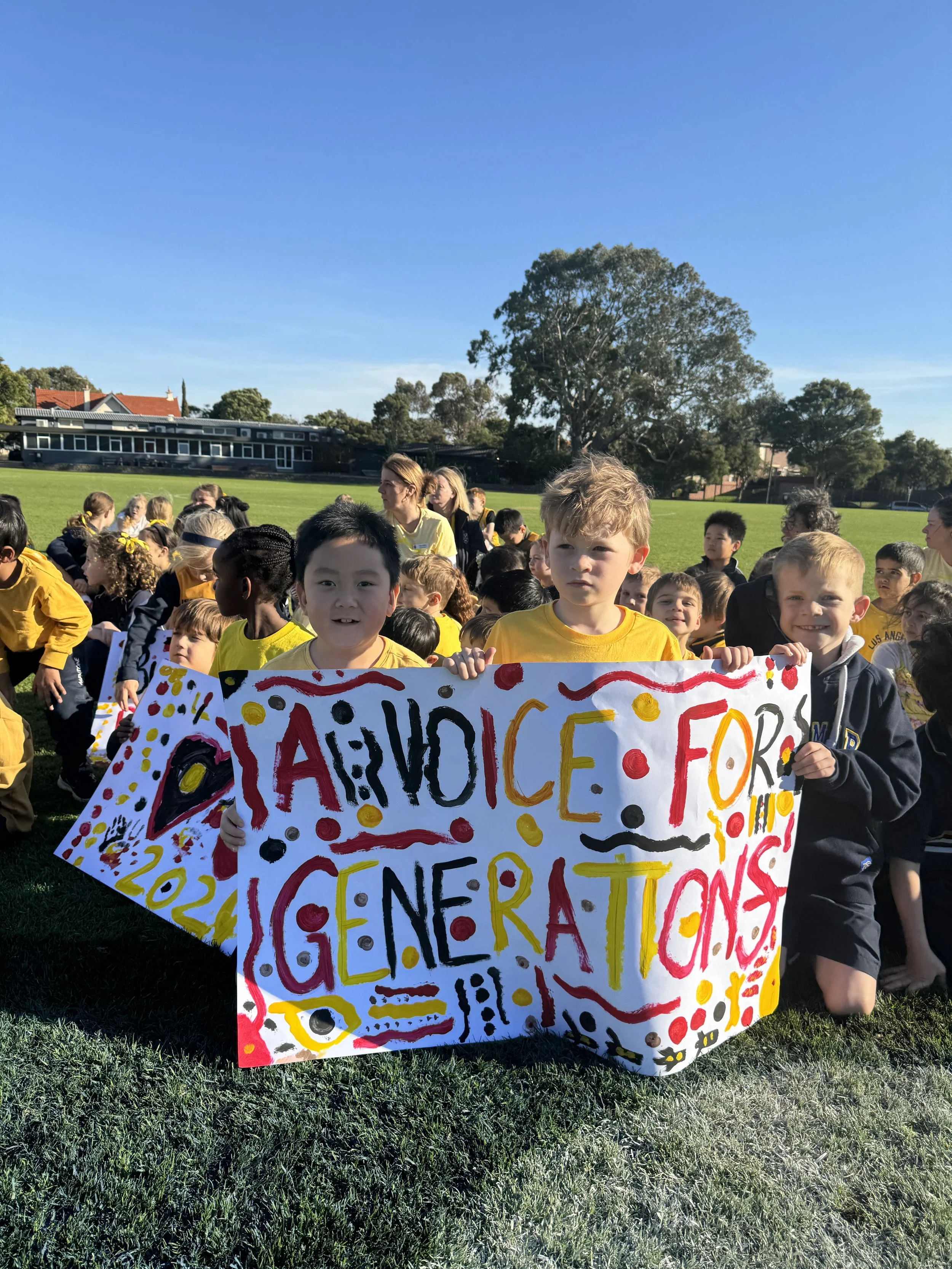 A group of children and adults gathered outside on a grassy field during daytime, holding a large, colorful handmade sign with the words 'Thank You for Your Science Celebrations' and various decorative patterns.