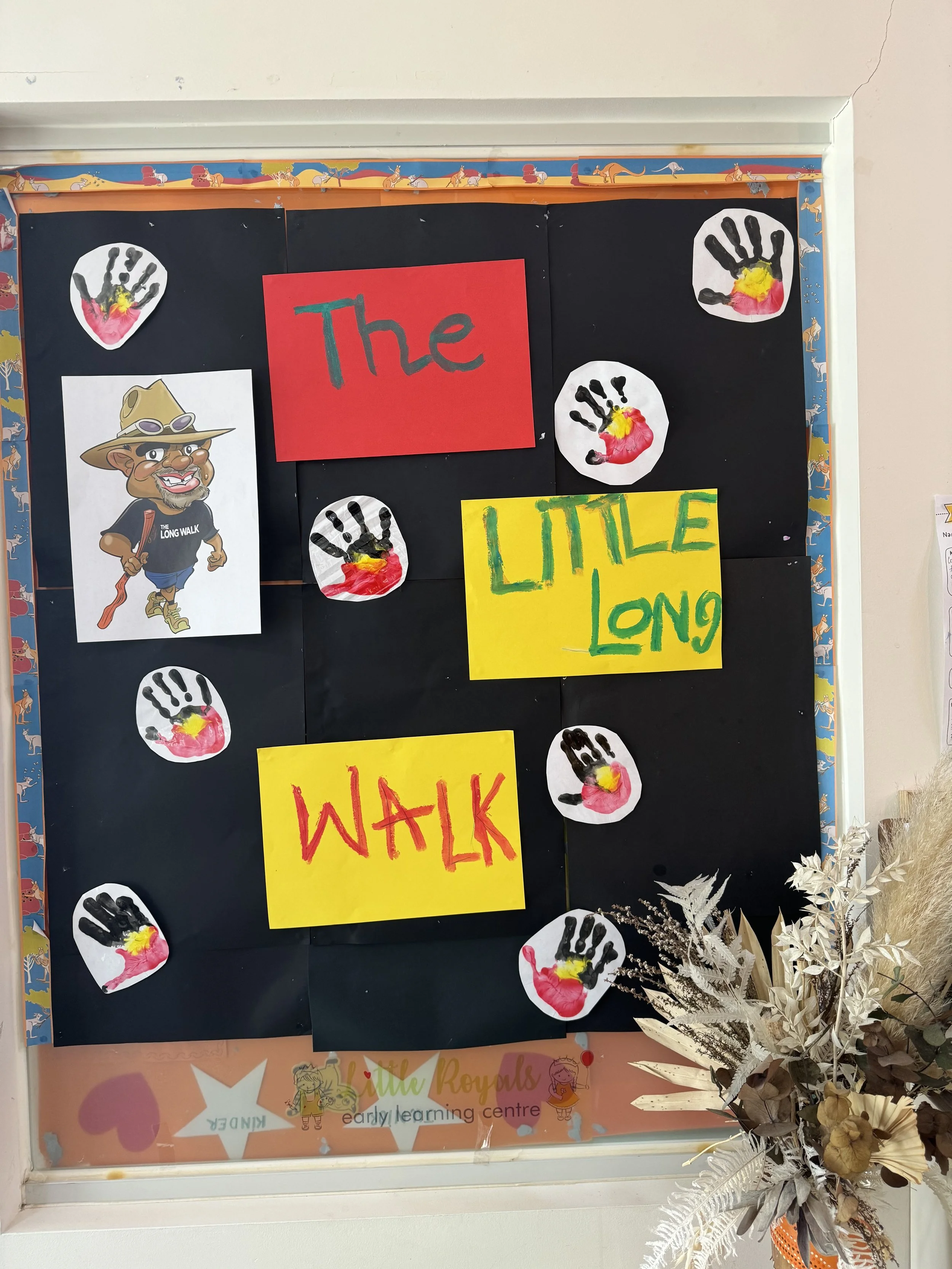 Decorative bulletin board with colorful signs and handprint art, featuring the words 'The' and 'Long Walk' and surrounded by paper flowers and foliage.