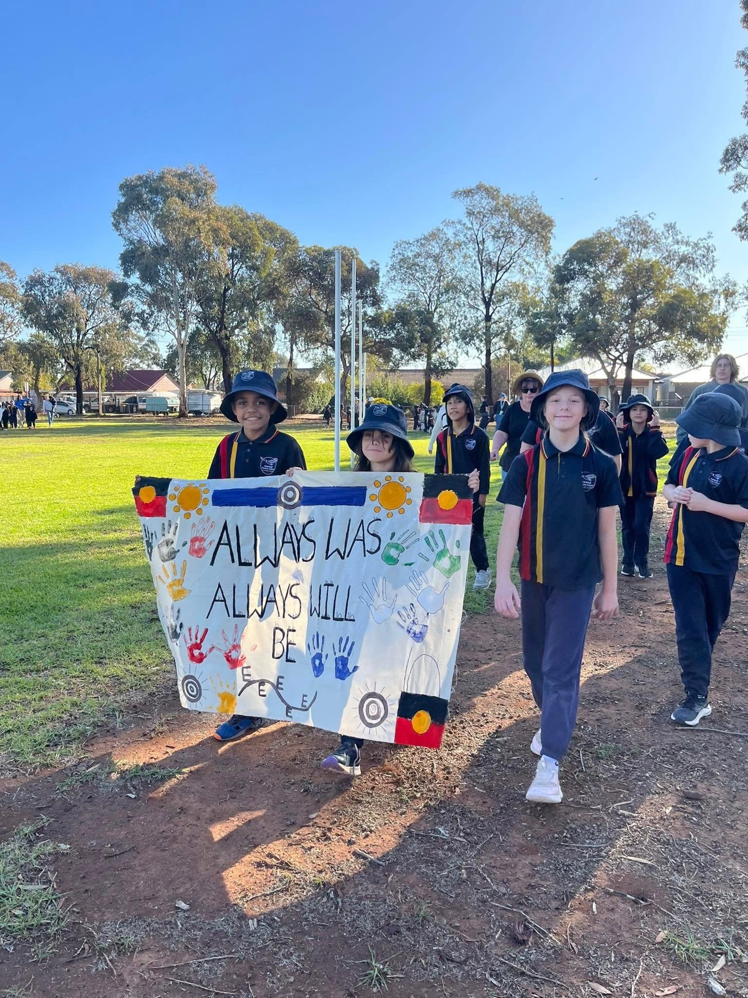 Children in school uniforms parade outdoors holding a colorful poster with handprints and the words 'All Ways Will Be Free' during a sunny day at a park or school grounds.
