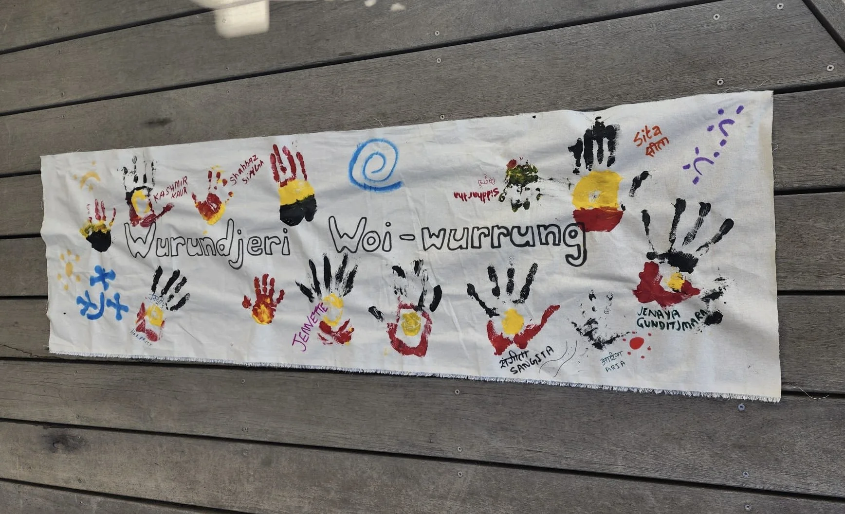 Colorful handprints in red, yellow, and black with names written beneath each, painted on a white piece of fabric laid on a wooden surface. Handprints include names like Kashmir Kaur, Shahnaz Singh, Jemaya Gunditjmara, and others. The central text re
